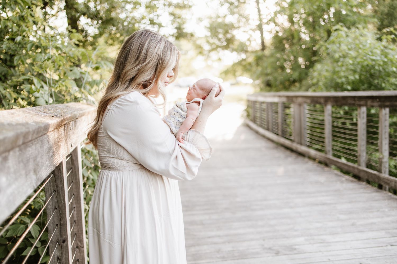 A mother in a cream dress holds her newborn baby on a bridge surrounded by lush green trees during lifestyle newborn photos in Lakeville, MN