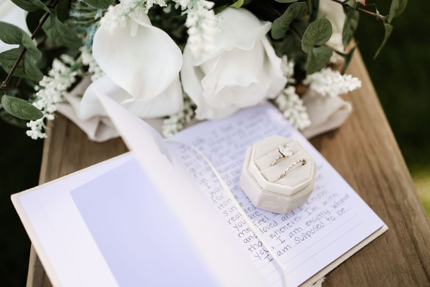 Wedding ring in white box on open journal beside white rose bouquet on rustic wooden bench.
