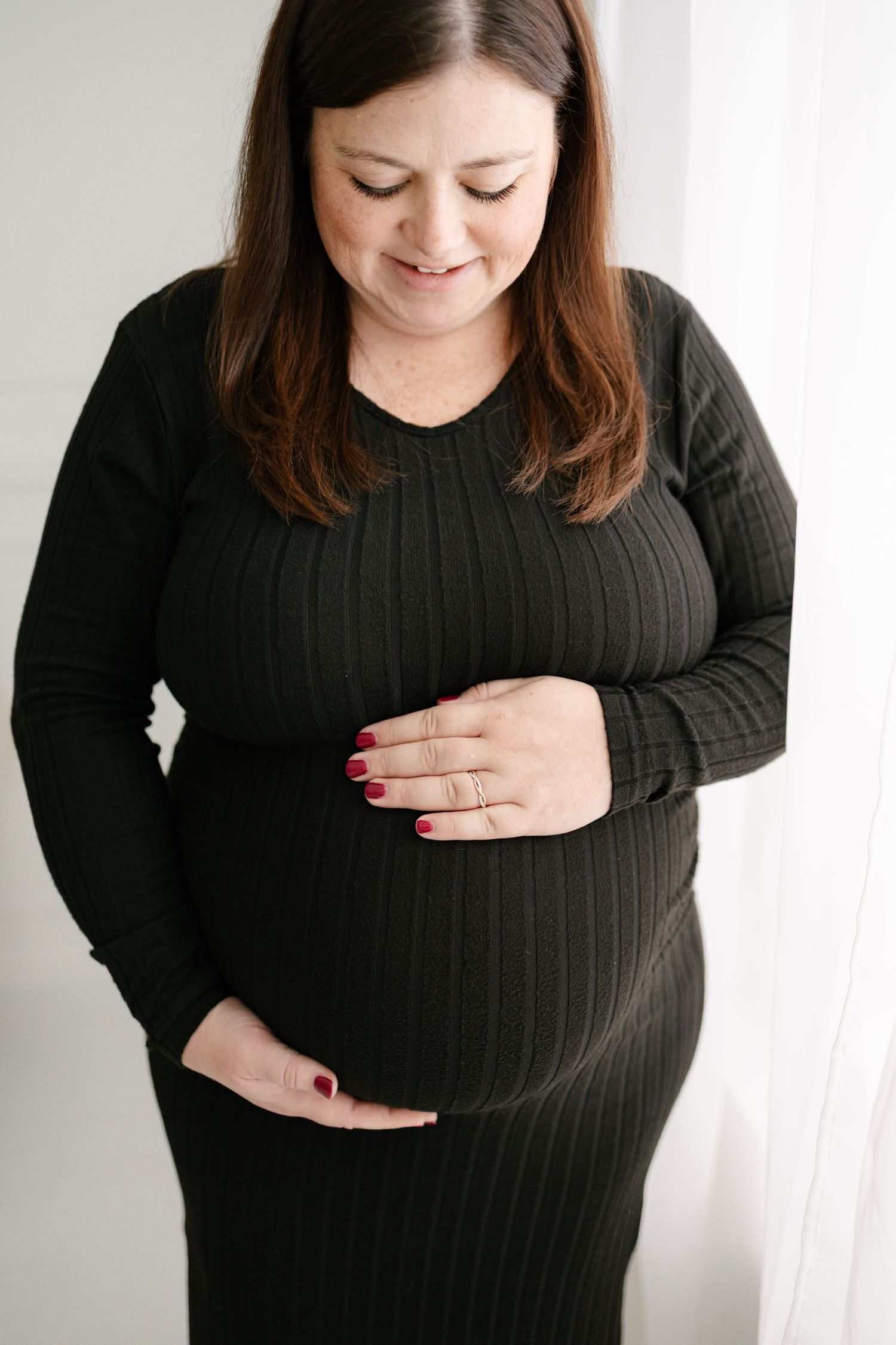 Smiling pregnant woman in black ribbed dress cradling her baby bump with red-painted nails and a ring taken by Woodbury photographer