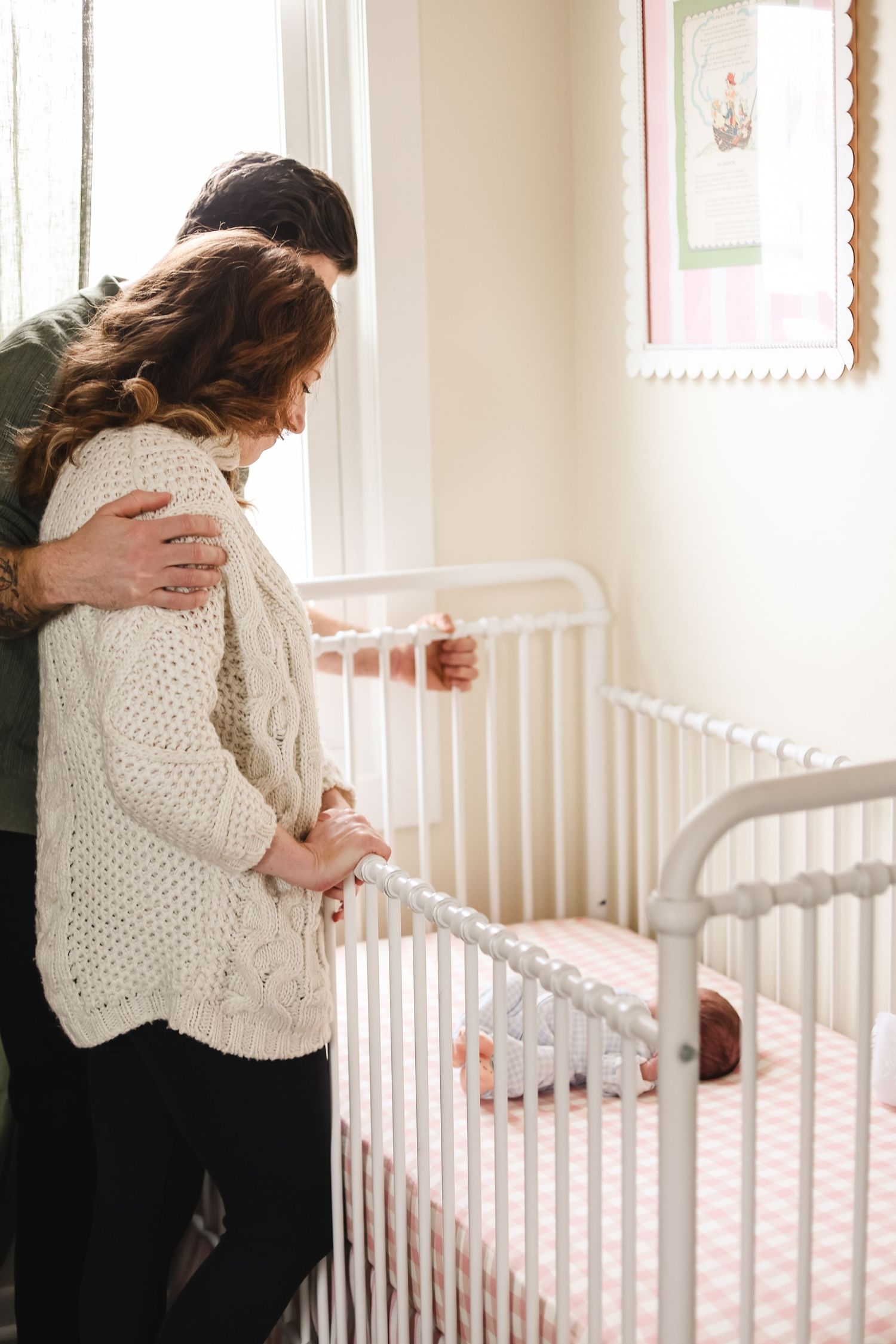 A couple stands lovingly by a white crib in a bright nursery room with a baby inside during Twin Cities in-home newborn session.