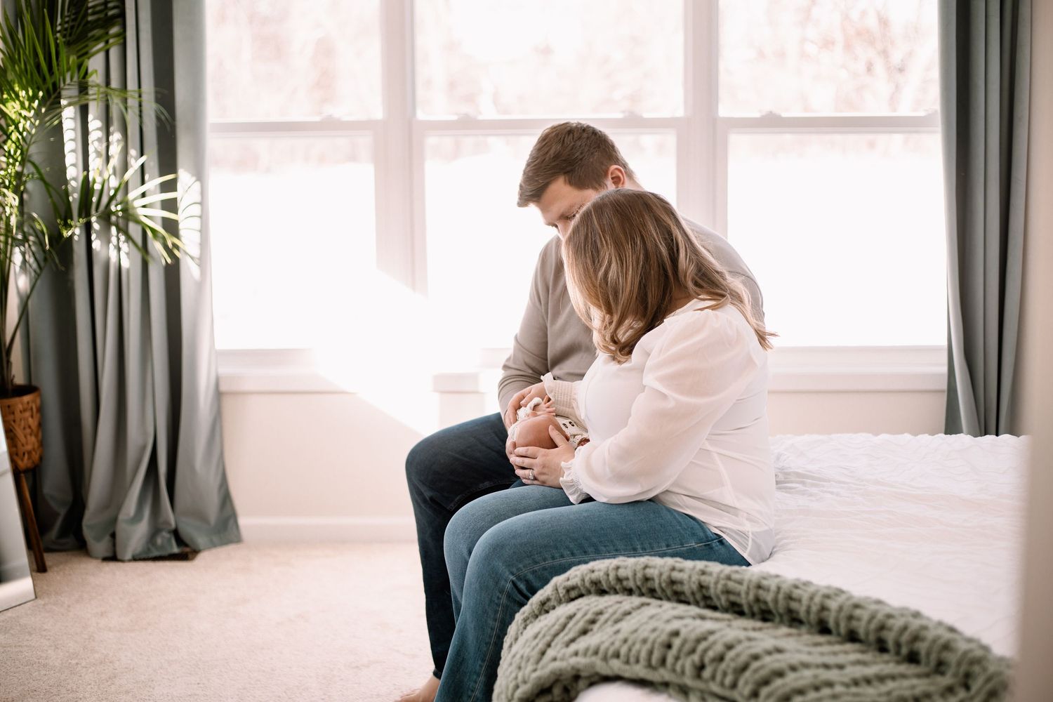 Parents sit close together cradling their newborn baby on a bed and a knit blanket in a bright room during Woodbury in-home newborn session.