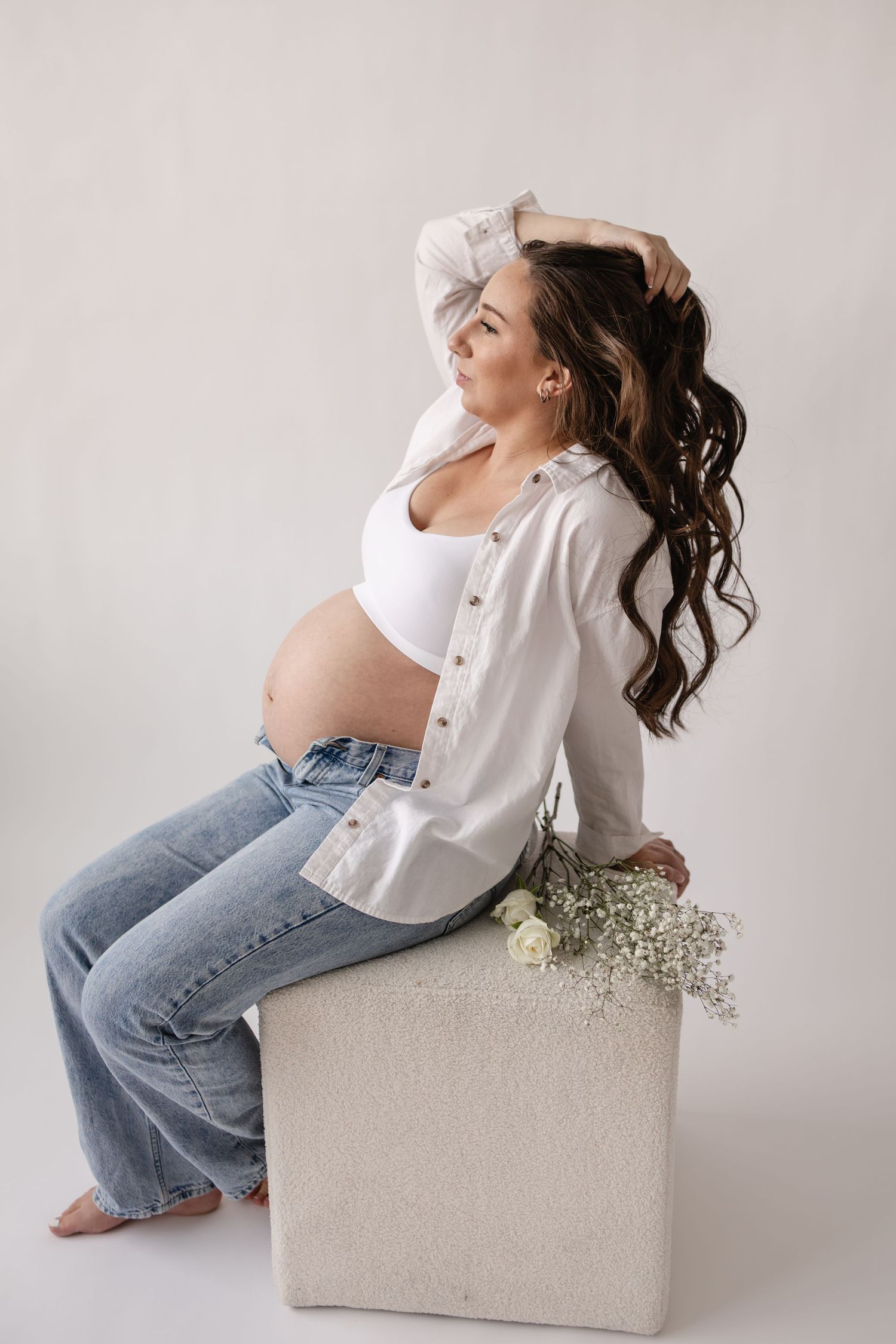 Artistic portrait series of a pregnant woman in white shirt and jeans posing on ottoman with baby's breath flowers in Minneapolis studio.