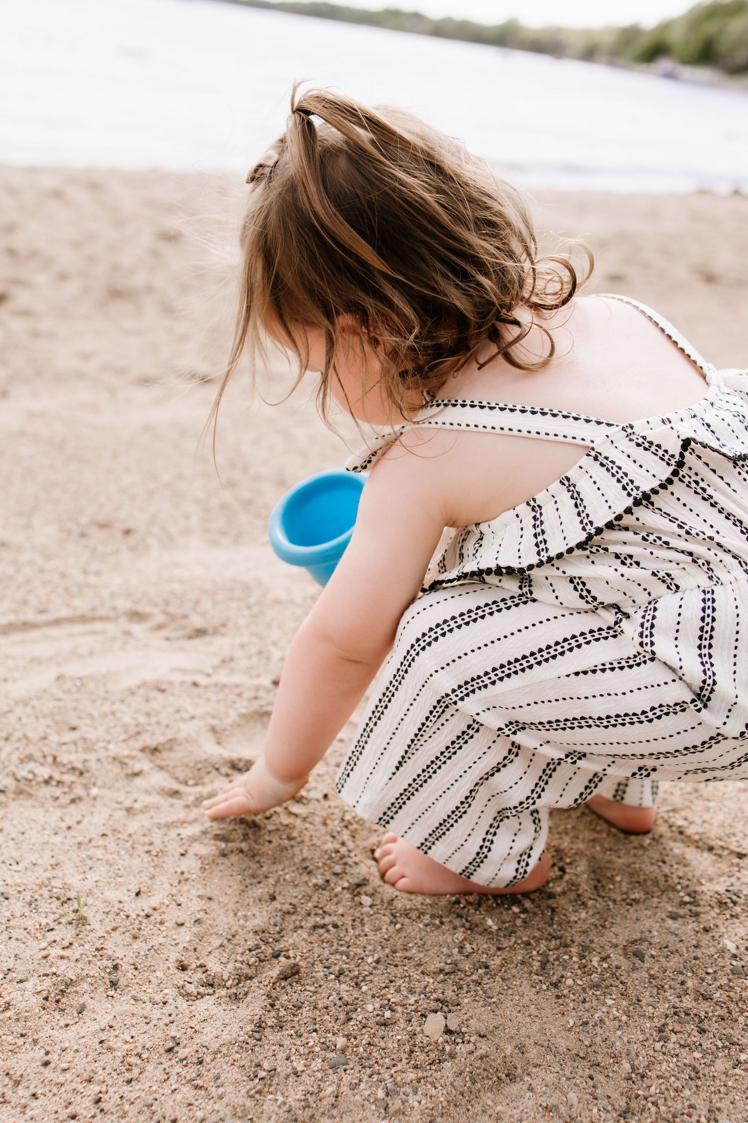 Young girl in striped dress playing in sand at beach, crouching down with blue bucket beside her.