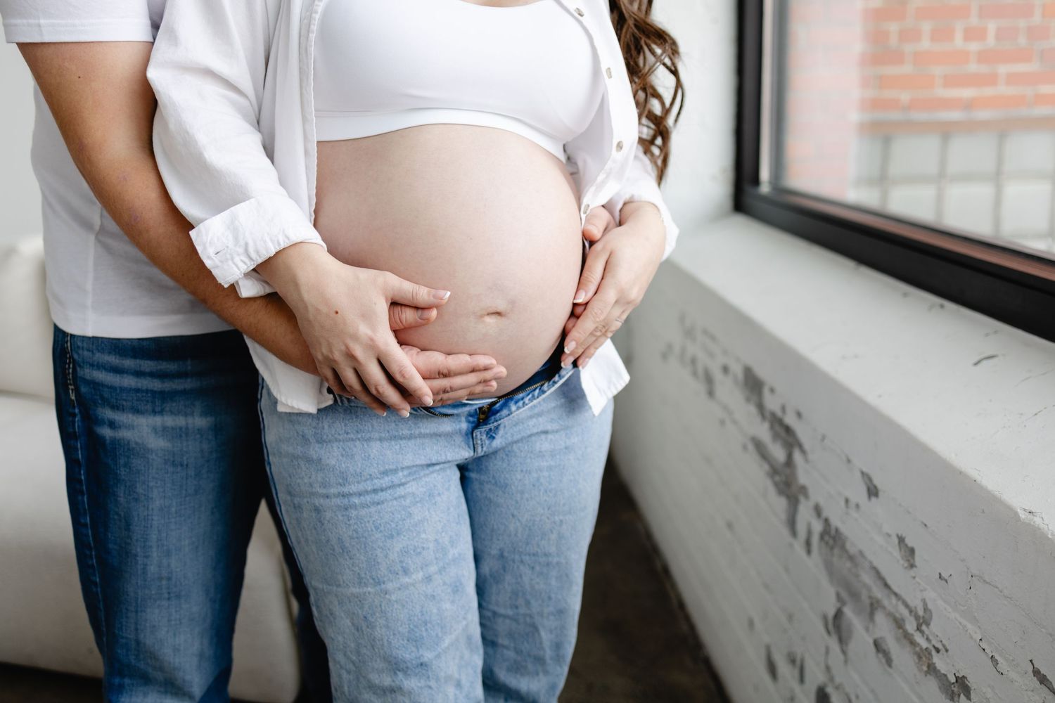 Pregnant woman in jeans cradling baby bump with partner's hands near window taken by Woodbury maternity photographer