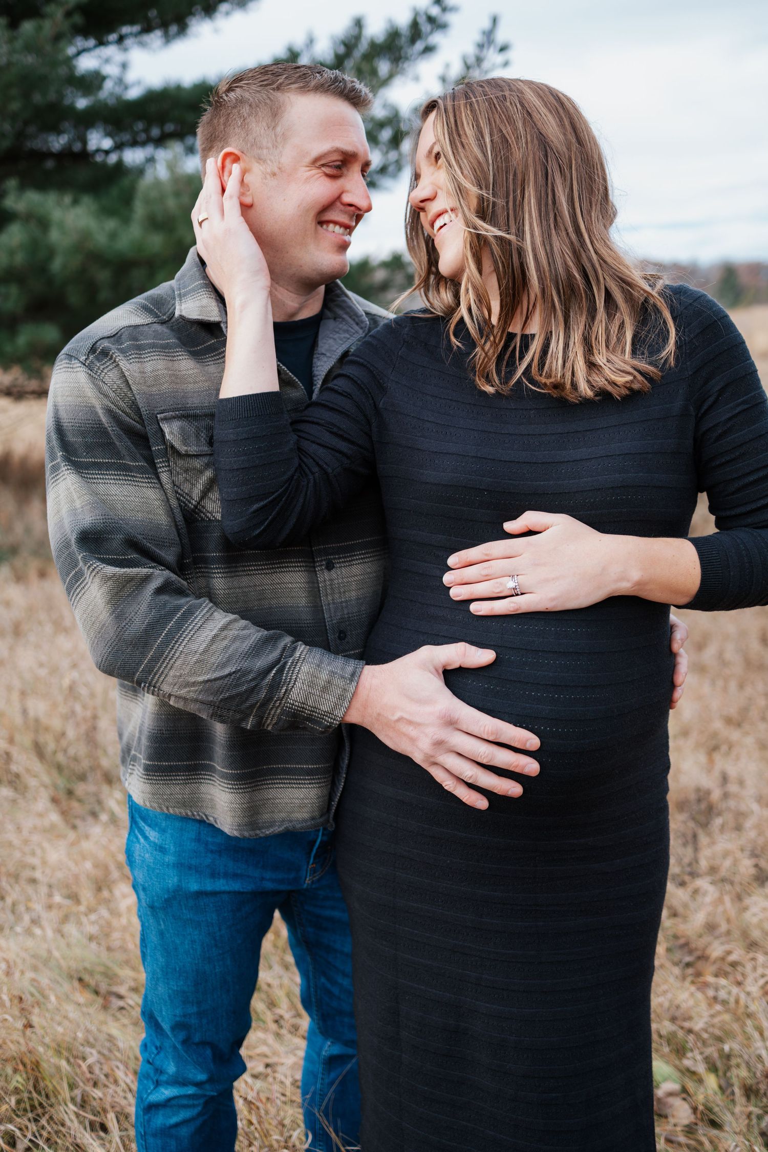 A pregnant couple in black and blue outfits standing in a field of tall grass on an overcast day, taken by Woodbury maternity photographer.