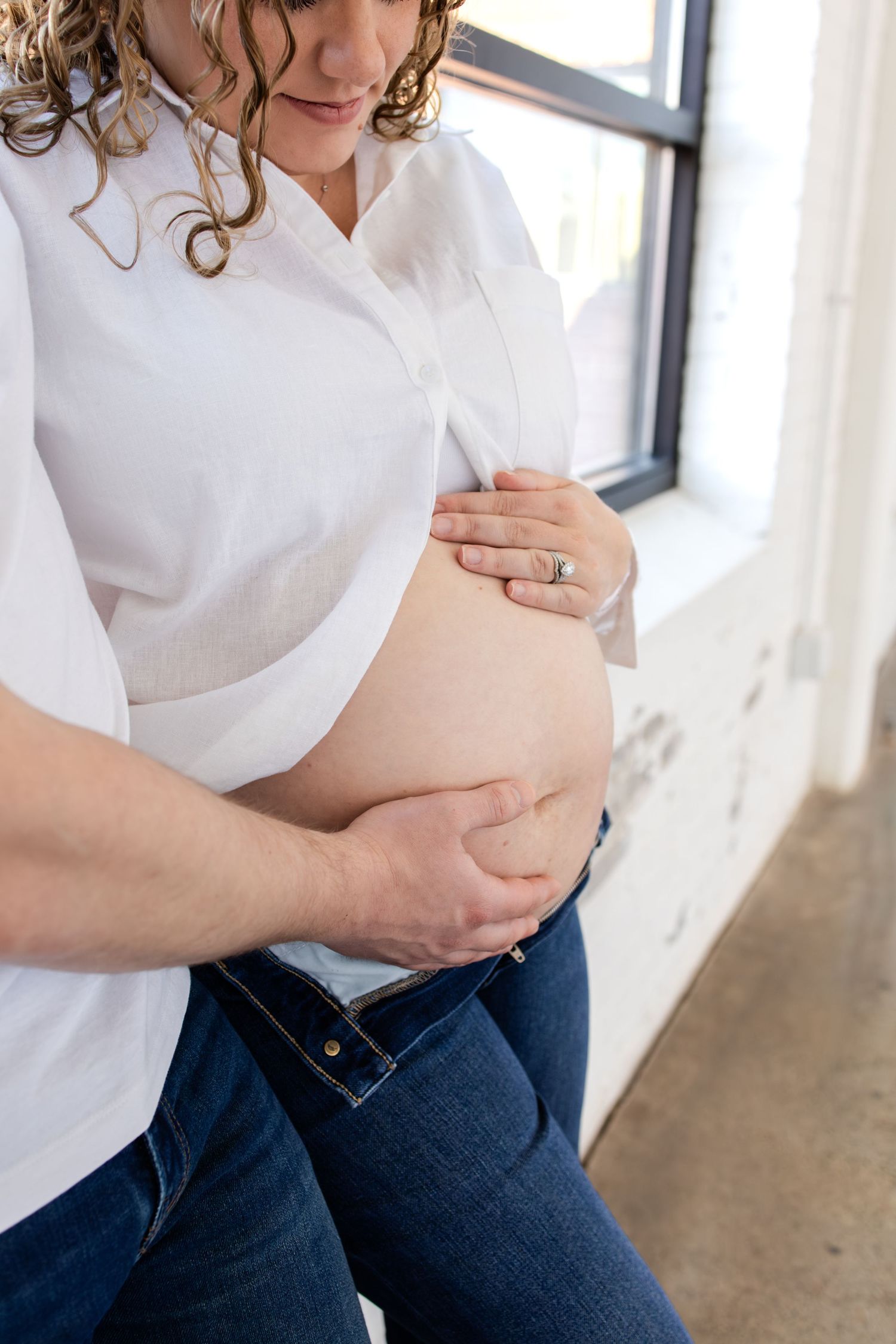 Pregnant woman in white shirt and jeans cradles baby bump while partner's hands lovingly support her belly near window in Minneapolis studio