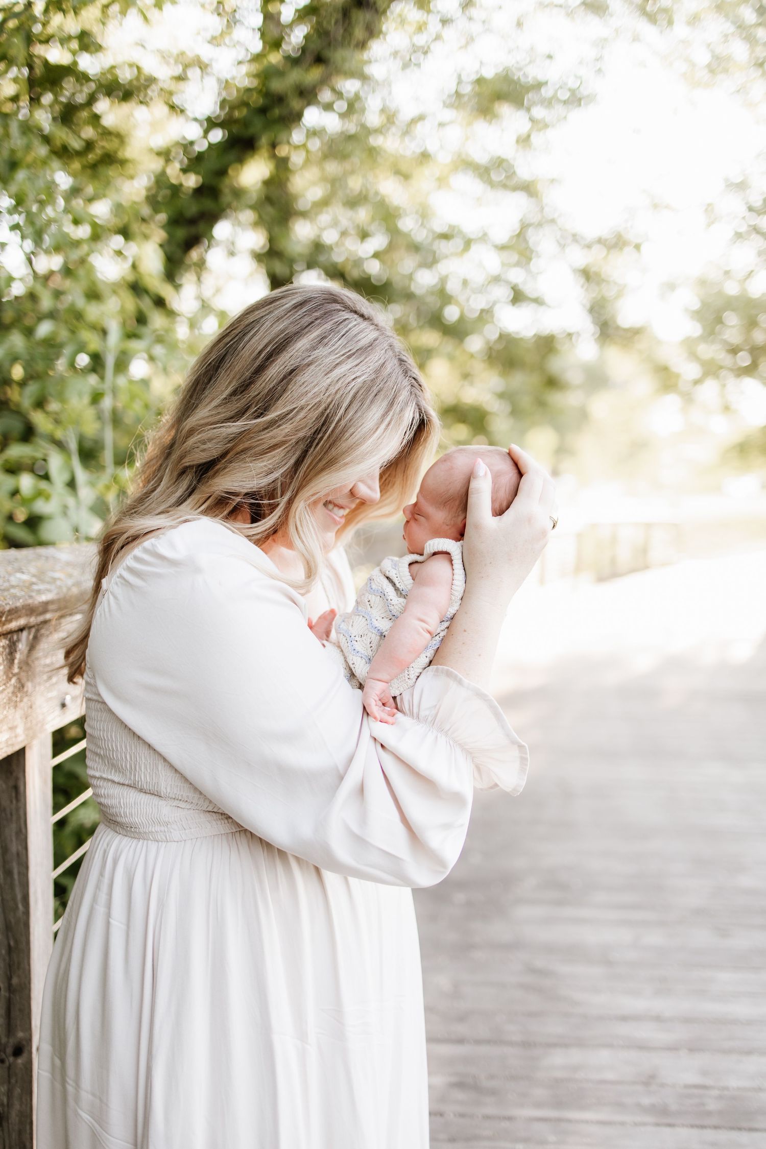 A mother in a white dress tenderly holds her newborn baby on a sunlit wooden bridge surrounded by green trees.
