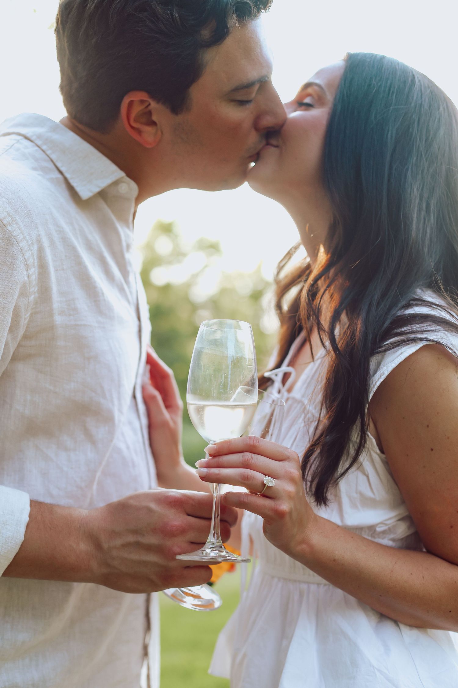 Romantic couple in white outfits sharing a tender kiss while holding wine glasses at sunset, taken by Woodbury engagement photographer.