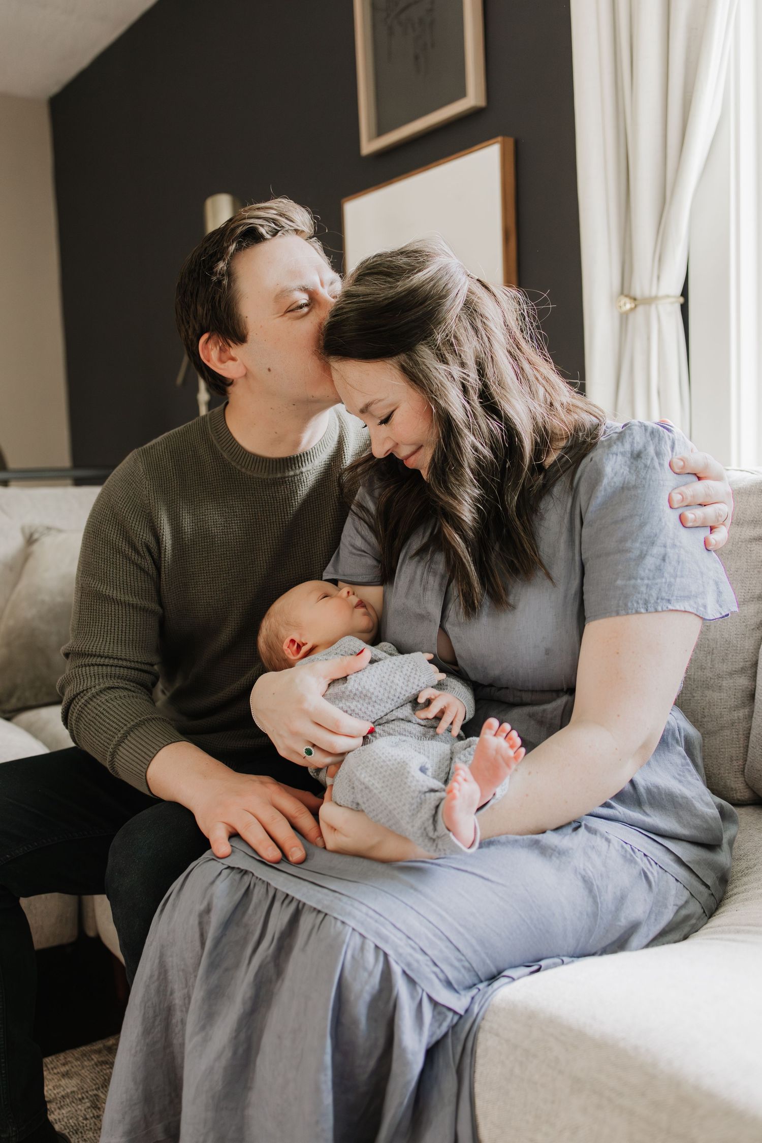 A couple sits on a couch, tenderly gazing at their newborn baby, sharing a loving family moment during in home newborn photos in Minneapolis