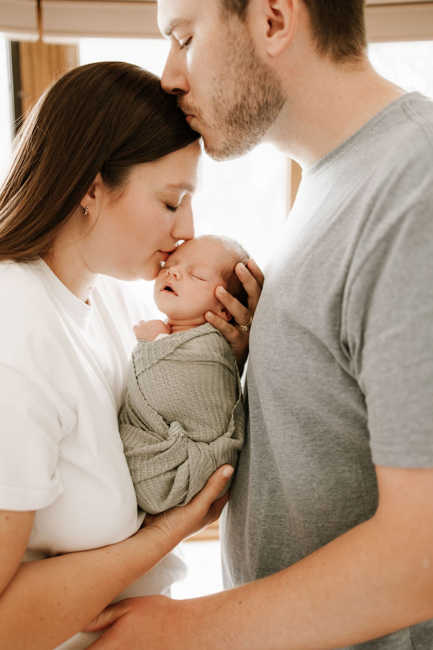 New parents tenderly kissing their sleeping newborn baby wrapped in a sage green swaddle blanket.