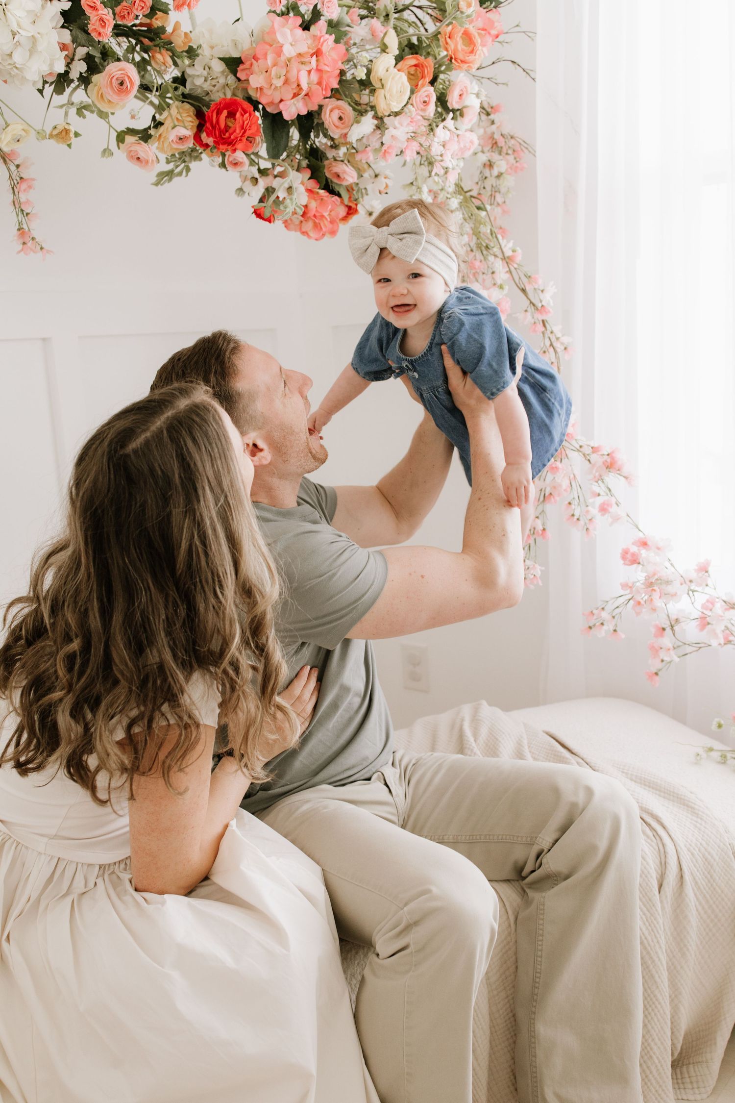 A couple sits on a bed lifting their smiling baby girl, surrounded by beautiful pink and white floral arrangements in Lakeville studio