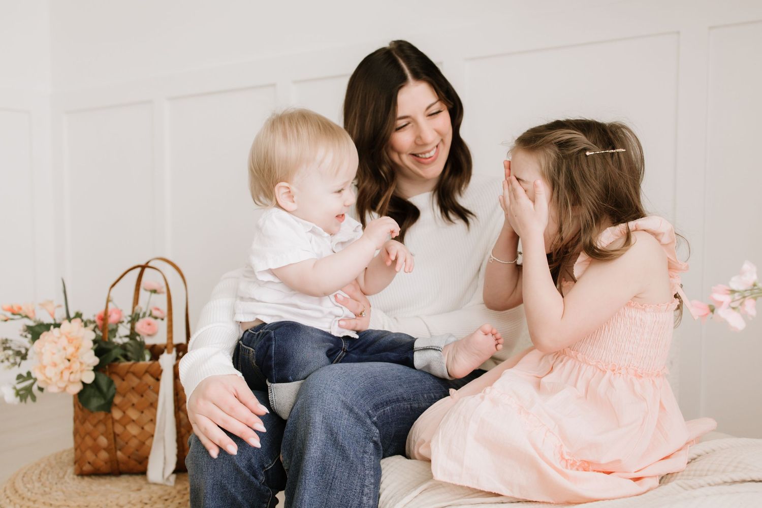 A mother sits with her baby and daughter in a pink dress in a bright white studio family photo session in Lakeville