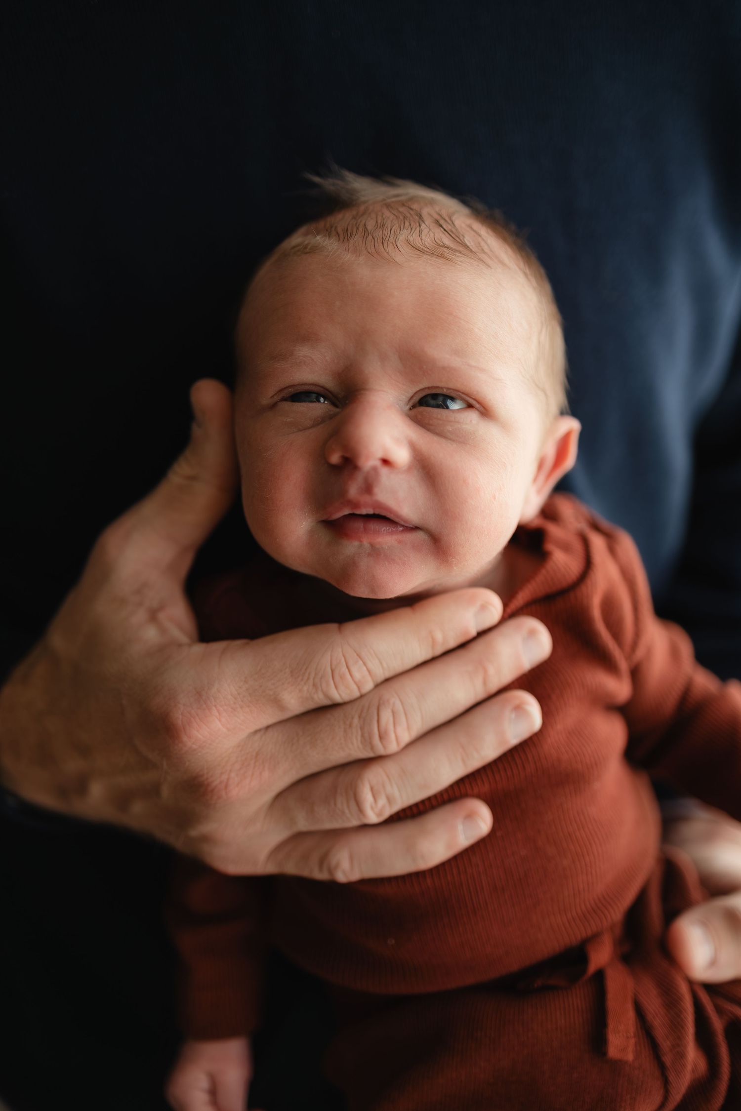 Newborn baby in a rust-colored onesie being cradled by gentle hands against a dark background during Twin Cities in-home newborn session.