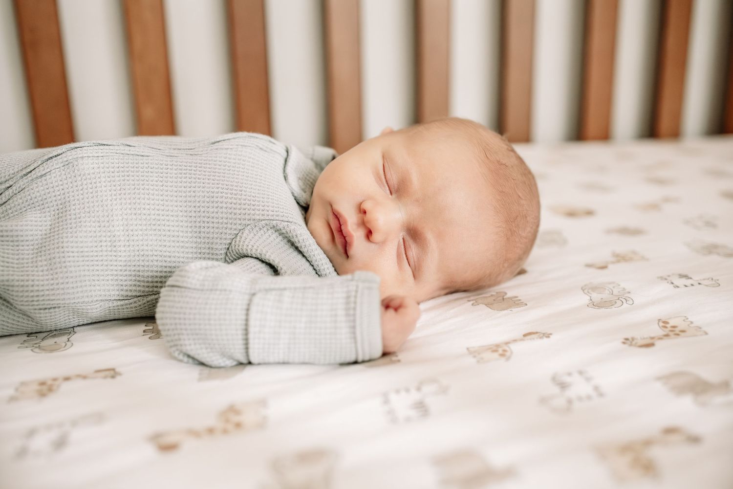 Peaceful newborn baby sleeping in a wooden crib wearing a light blue waffle knit sleeper on patterned sheets during Woodbury newborn photos