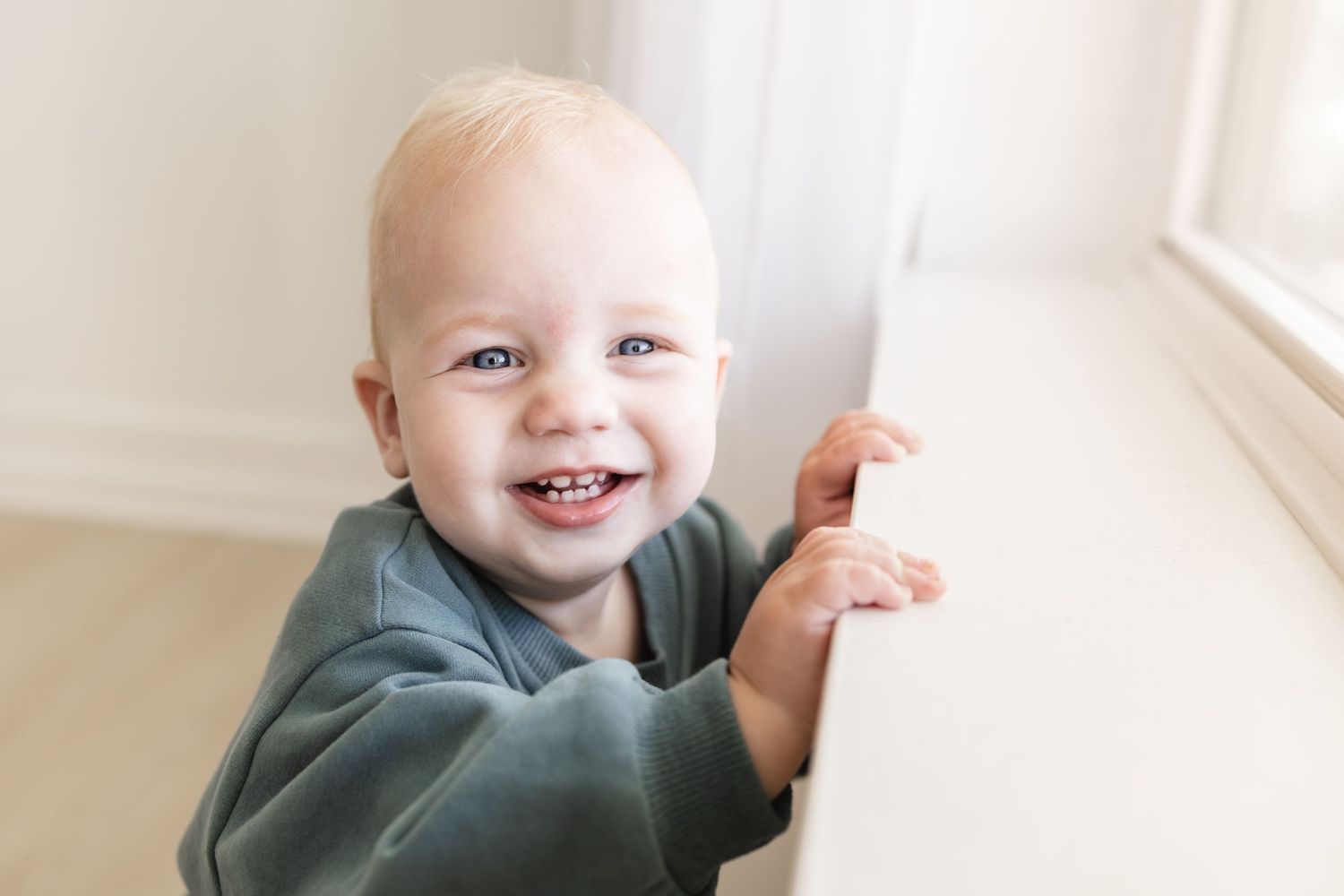 Smiling baby with blue eyes standing at a white crib rail in a bright nursery room.