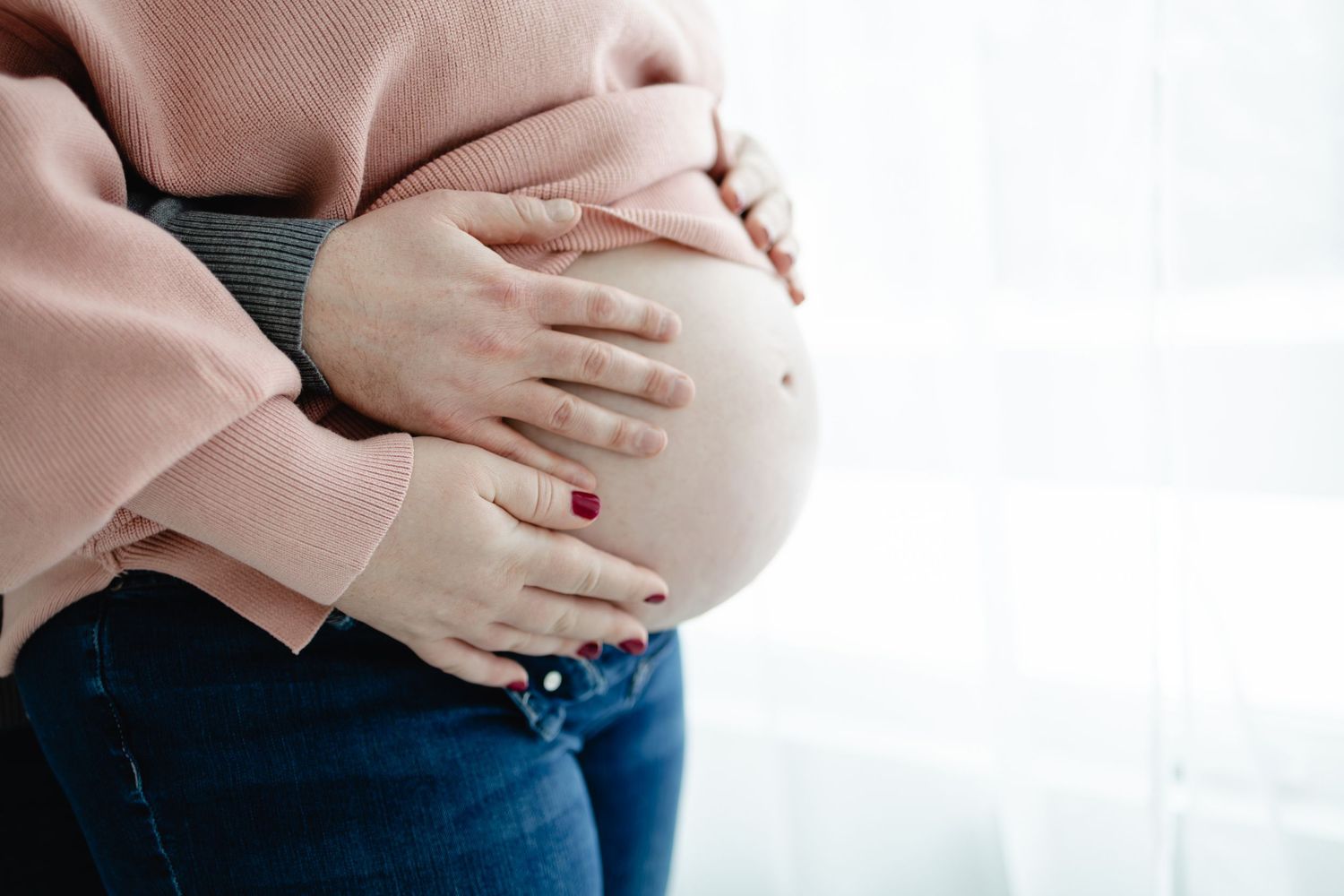 Pregnant woman in pink sweater and jeans cradling her baby bump with both hands taken by Woodbury maternity photographer