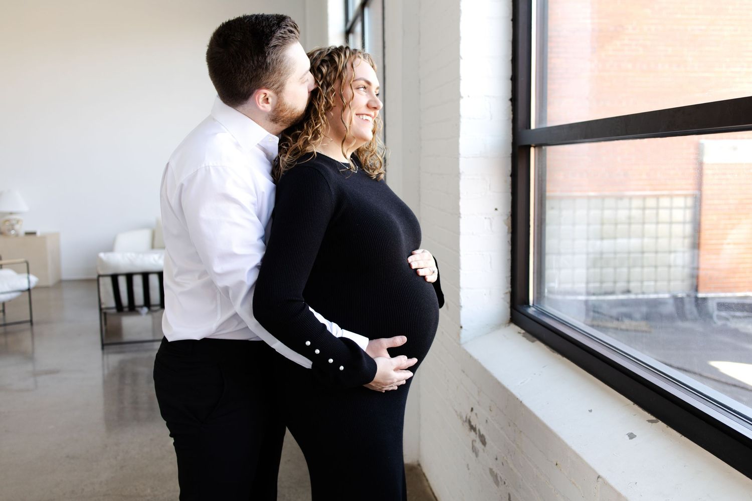 Couple sharing a tender maternity moment by a window, woman in black dress cradling baby bump taken by Minneapolis maternity photographer