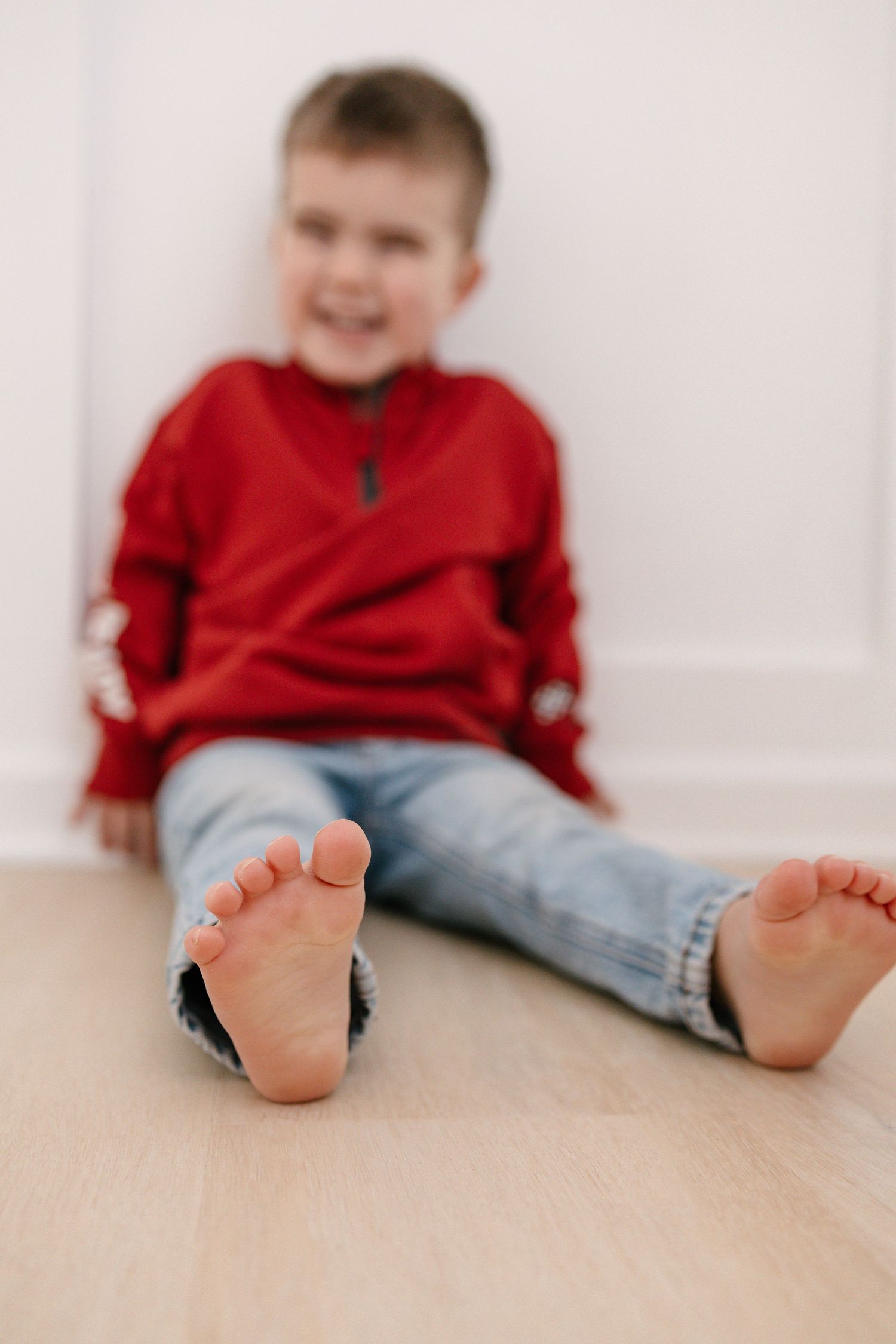 A smiling young boy in a red sweatshirt and jeans sits barefoot on a light wood floor against a white wall in Lakeville studio