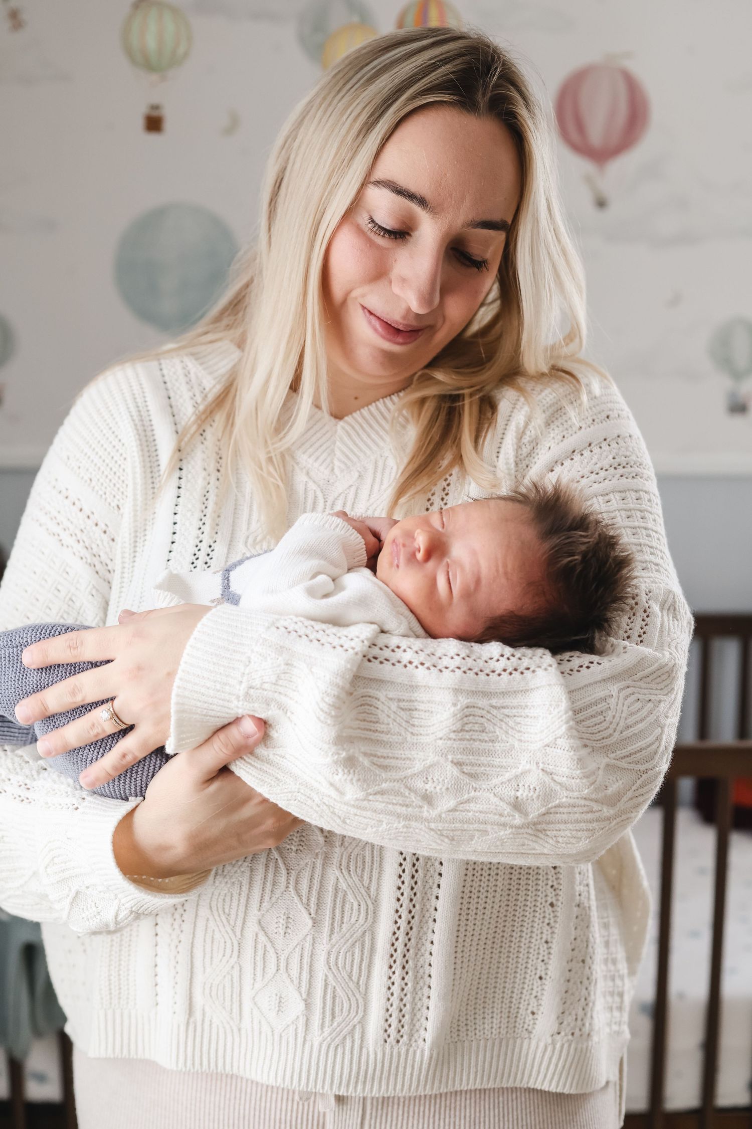 A mother wearing a white sweater holds and gazes lovingly at a newborn baby in a bright, airy room in in-home Twin Cities newborn session.