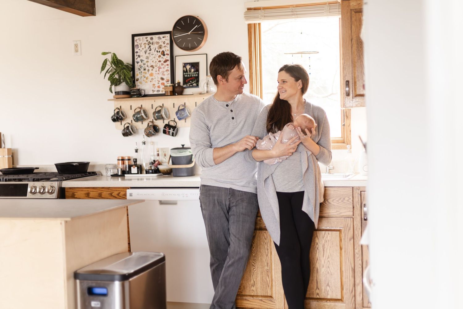 Two parents chat and laugh together while they cradle their newborn in a bright modern kitchen space during Afton newborn photo session.