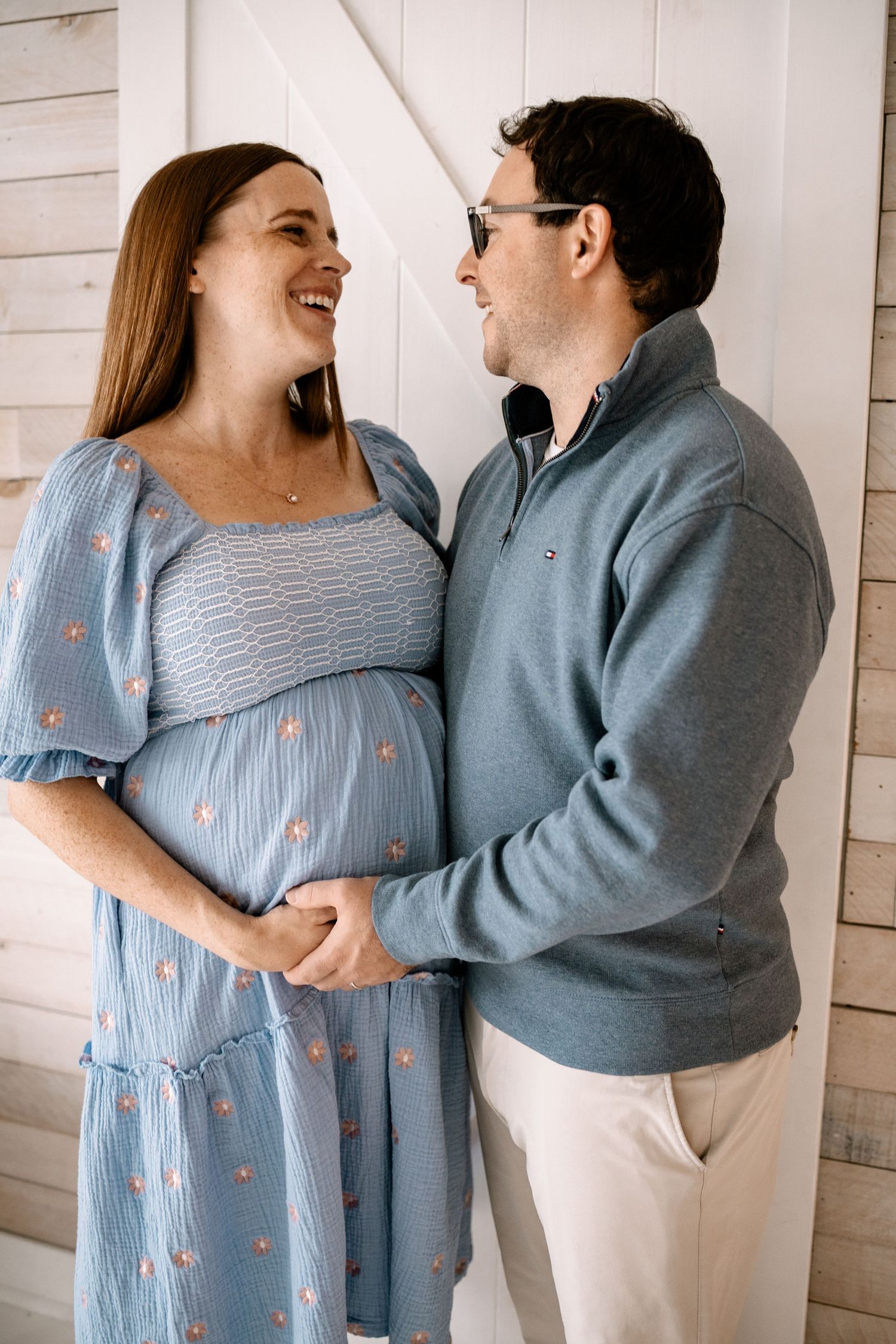 Happy couple sharing a tender moment, pregnant woman in blue floral dress held by partner in gray sweater during Woodbury maternity session