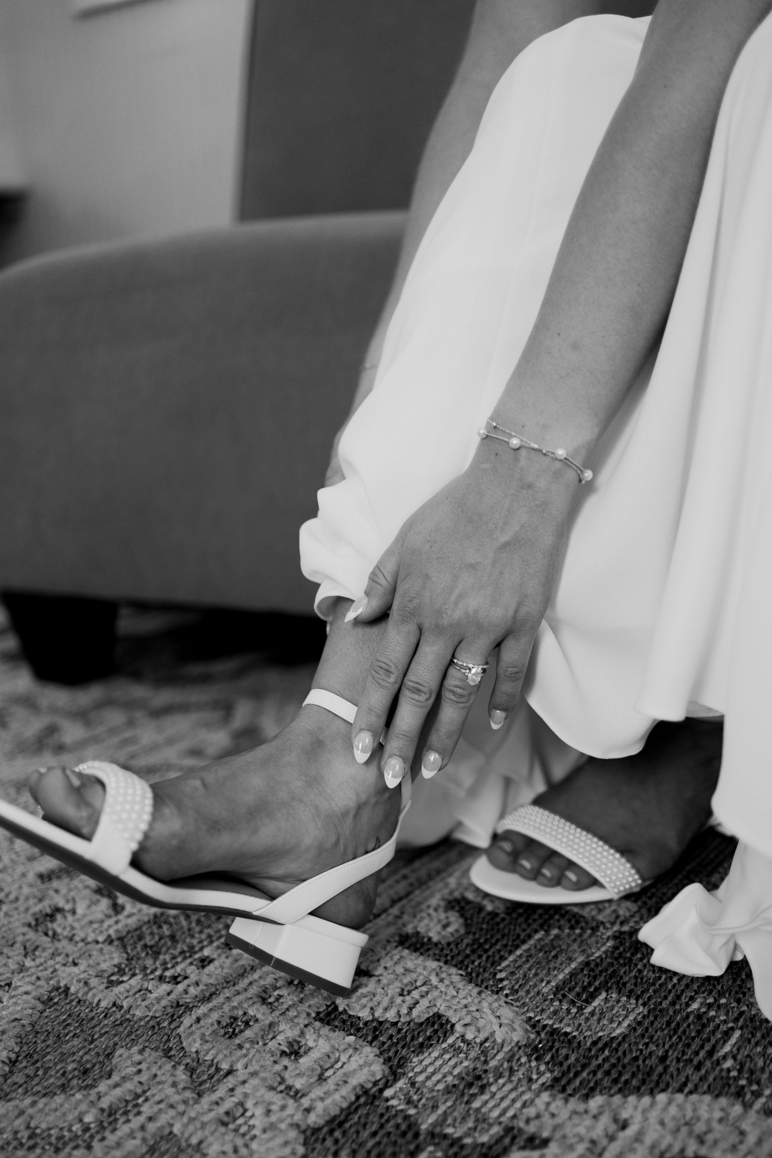 Black and white photo of a bride putting on white heeled sandals, wearing a ring and bracelet, with white dress.
