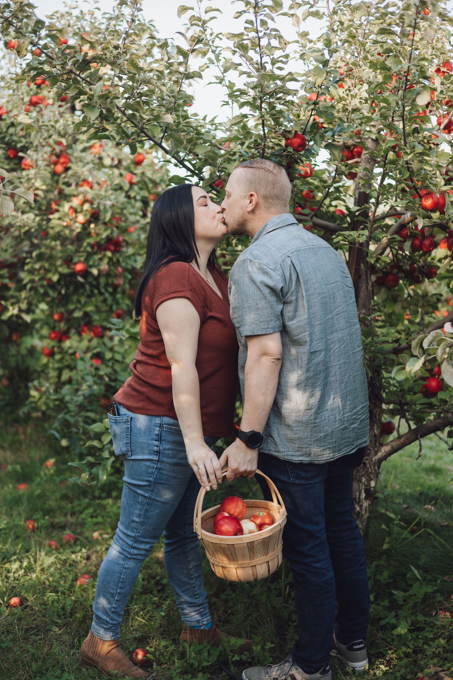 A couple shares a romantic moment while apple picking in an orchard holding a woven basket, taken by Lakeville couples photographer.
