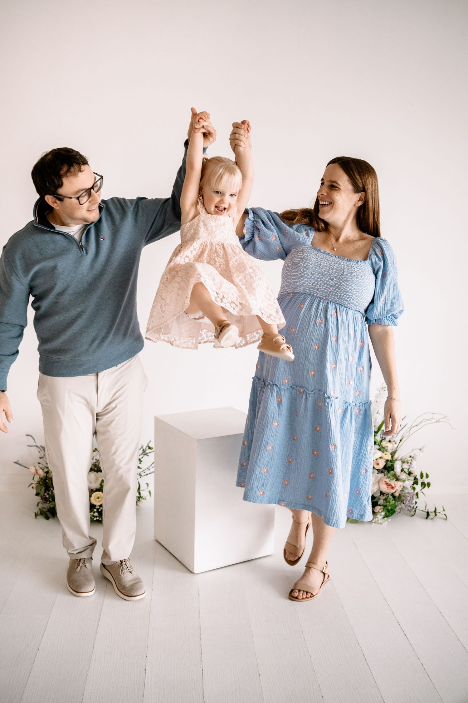 Happy family with parents lifting smiling baby girl in pink dress during a bright studio photo session in Farmington.