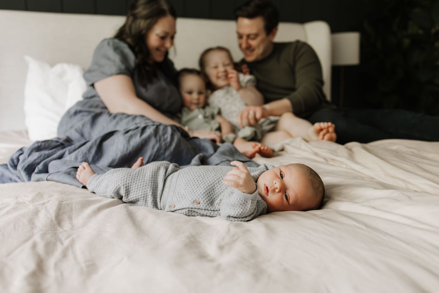 A newborn baby lies on a white bed while a happy family with two toddlers smiles together in the background.