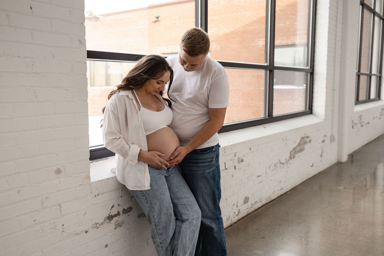 A couple in casual clothing embraces her pregnant belly lovingly in a bright white industrial loft space with large windows in Minneapolis.