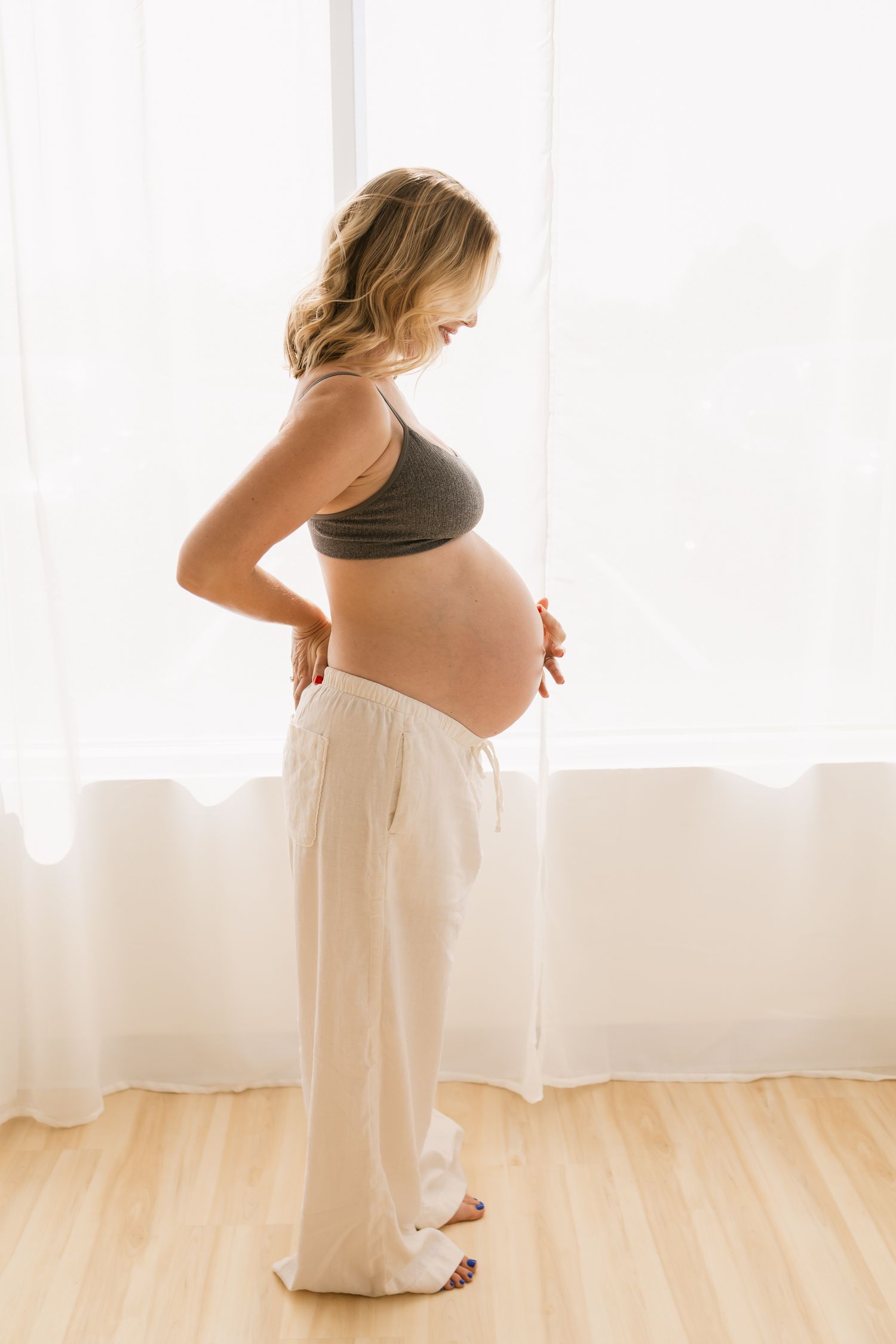 Pregnant woman in gray crop top and white pants cradling her baby bump while standing by a bright window.