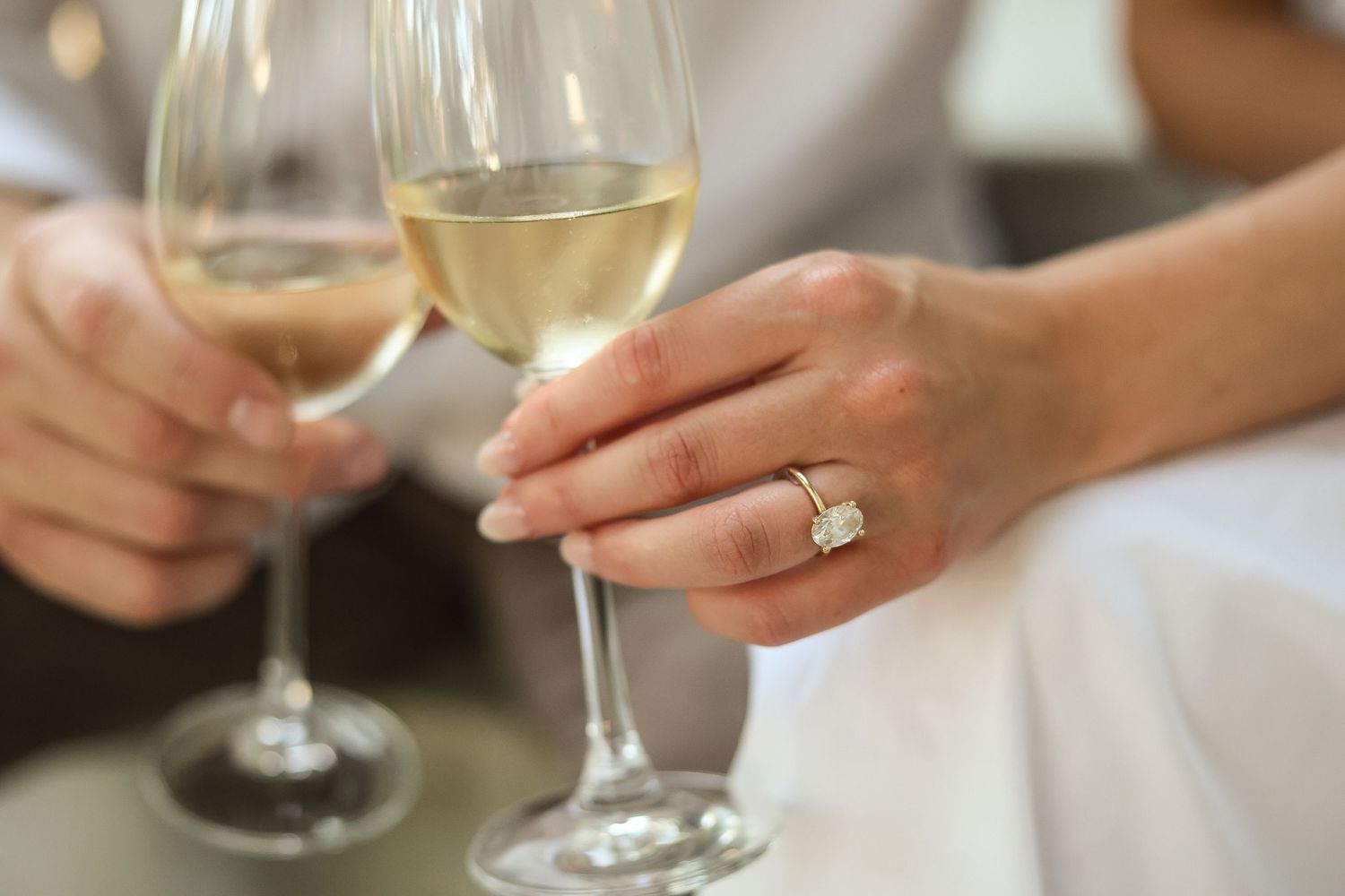 Two hands clink white wine glasses together in a celebratory toast while wearing an elegant ring during Woodbury engagement photo session.