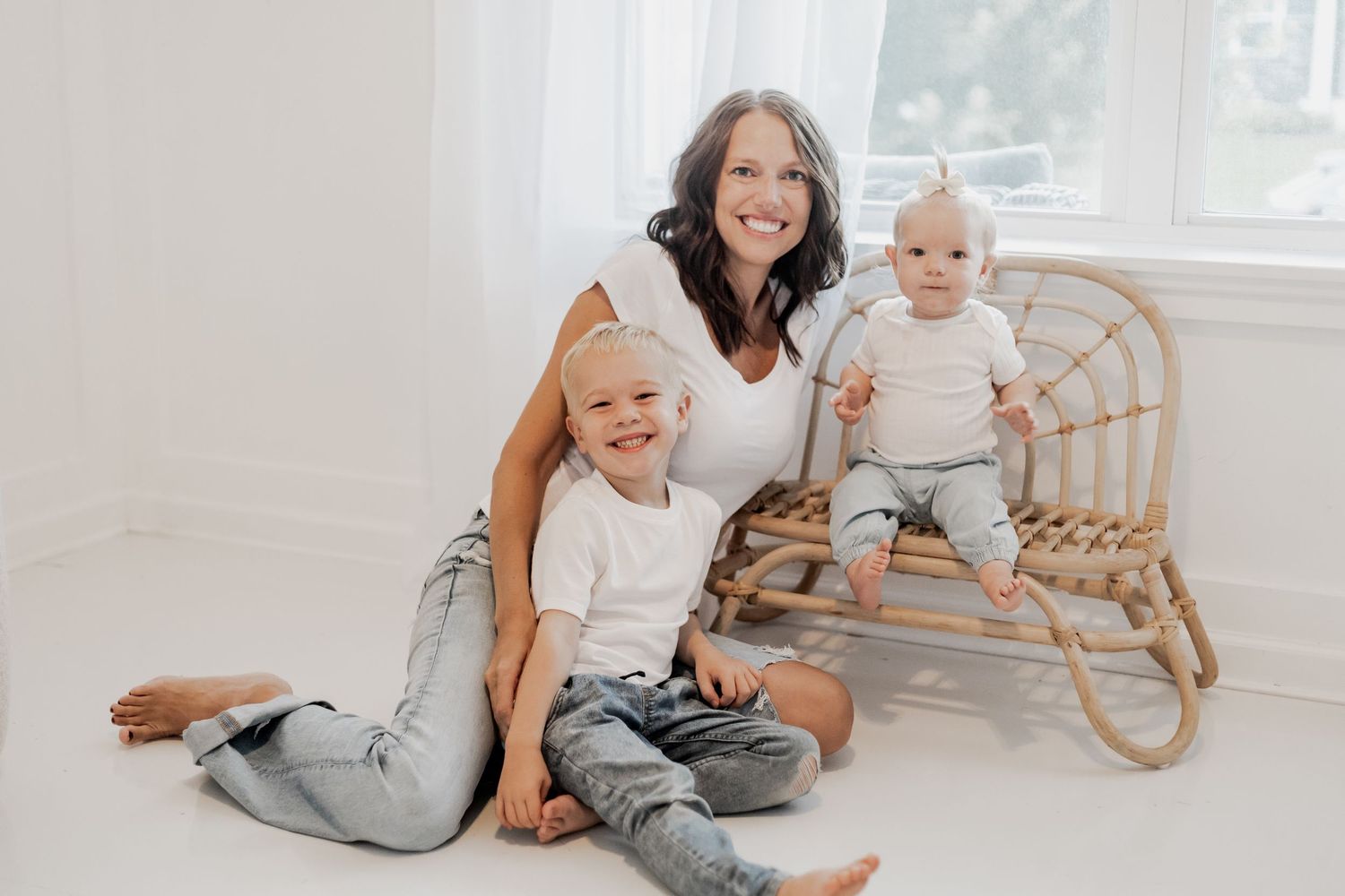 A family in coordinating white and denim outfits sits together on a rattan chair in a bright white room during Lakeville mini session.