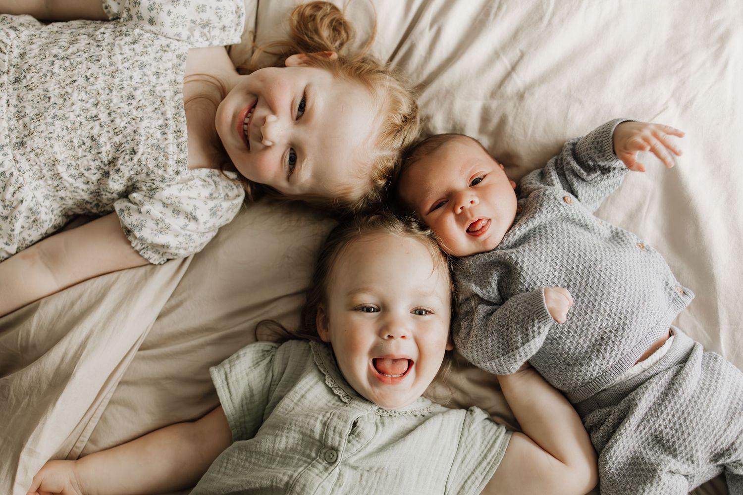 Three young siblings lying together on a white bed, smiling and laughing during in-home newborn session in Twin Cities