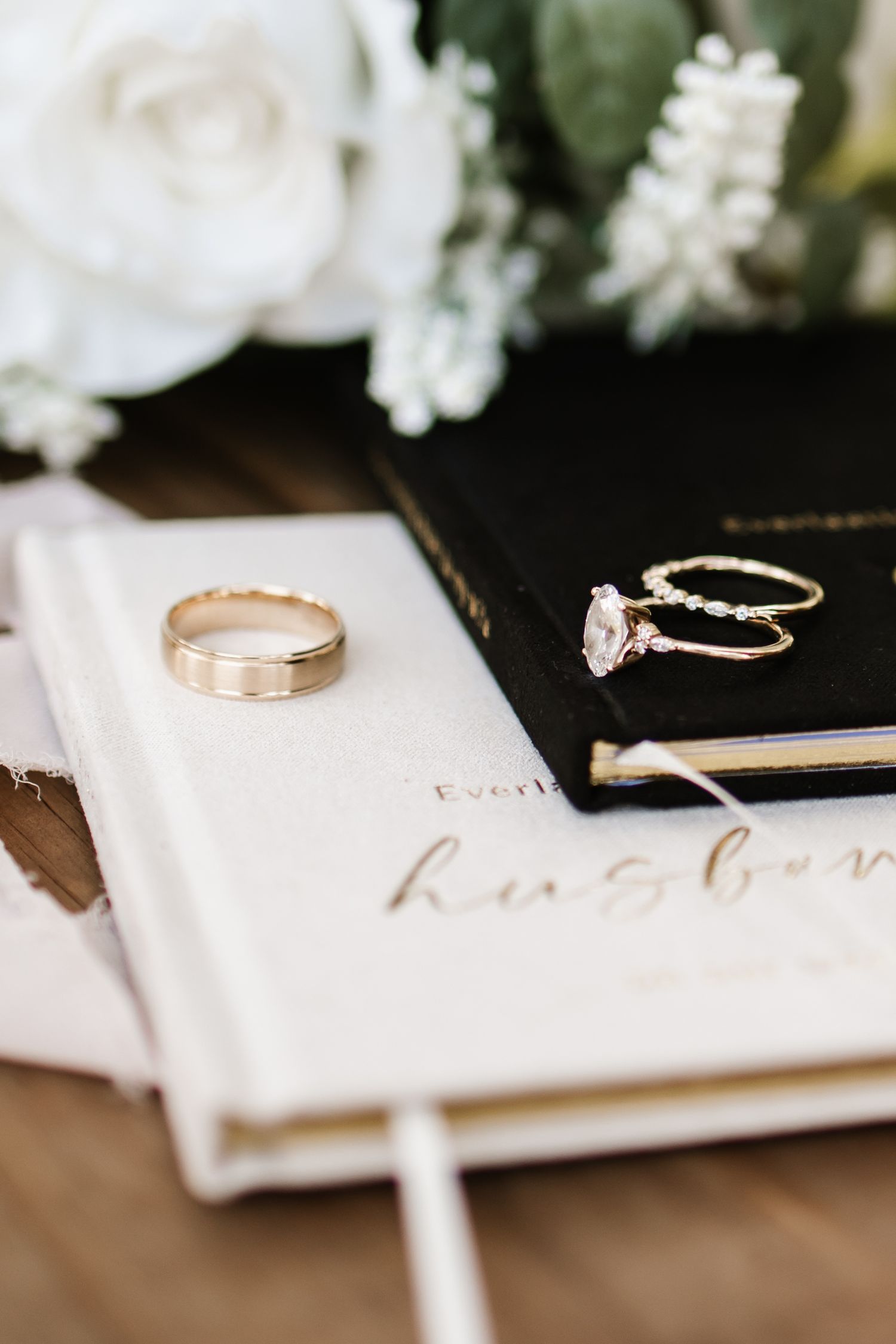 Gold wedding band and diamond engagement ring displayed on elegant white journal beside black book with white roses.