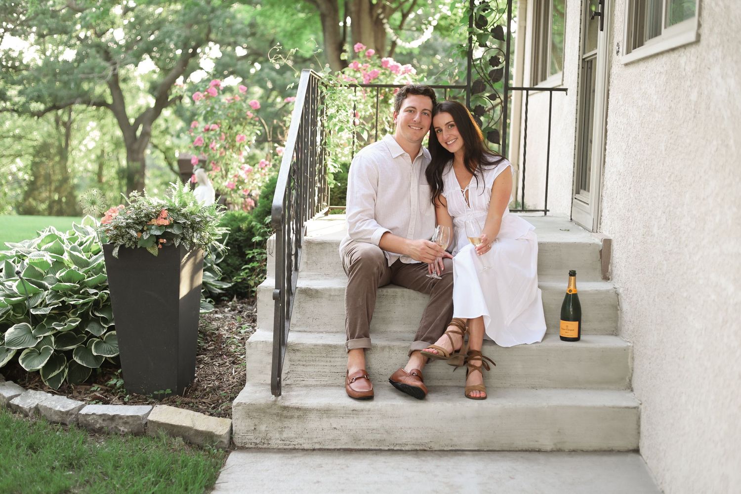 A couple sits together on a concrete porch step surrounded by greenery on a sunny day during Woodbury engagement photo session.
