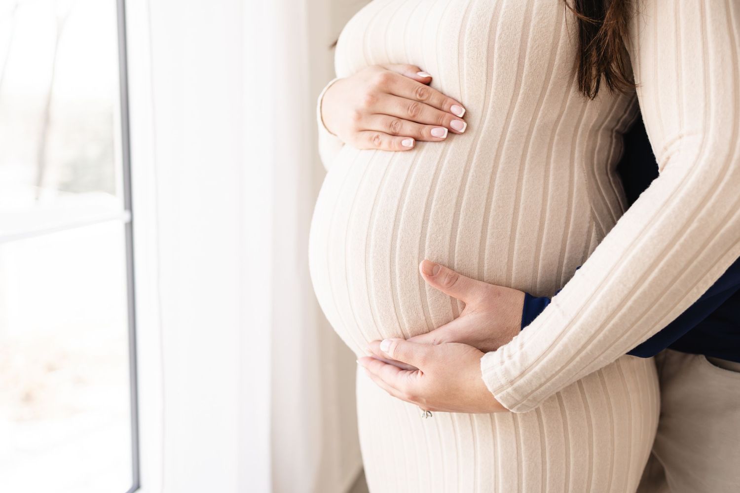 Couple embracing pregnant belly in cream ribbed maternity dress near bright window during Twin Cities maternity photo session
