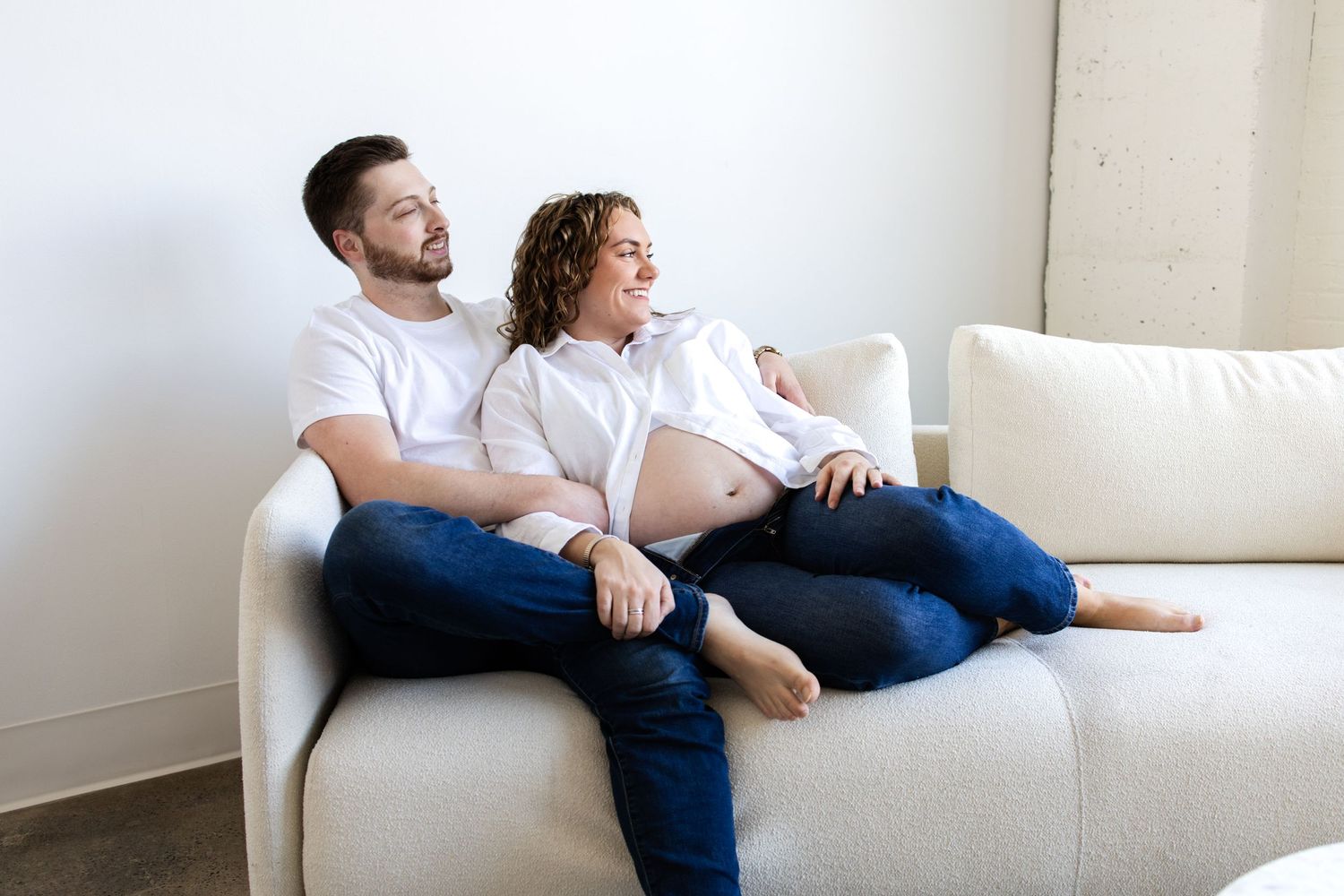 Pregnant couple sitting on a white sofa, smiling and relaxing together in a bright modern studio in Minneapolis maternity session