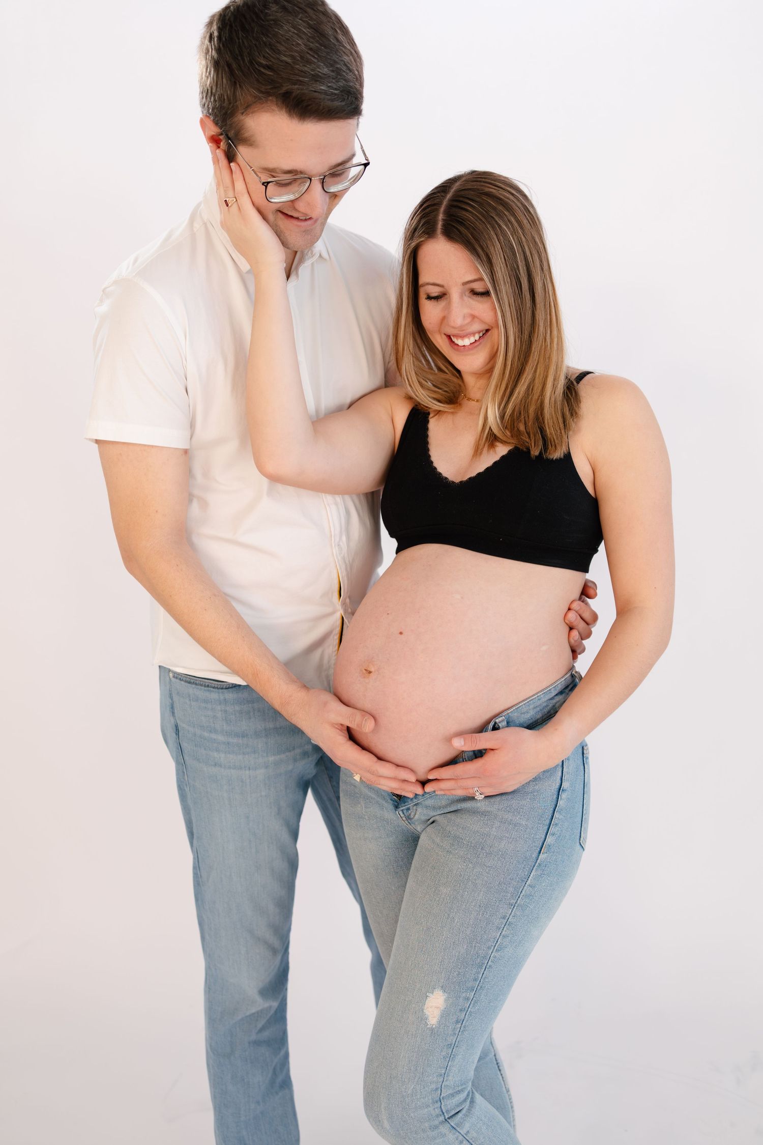 A happy couple posing for a maternity photo, with the man embracing the pregnant woman as both look down at her bare baby bump in Chaska