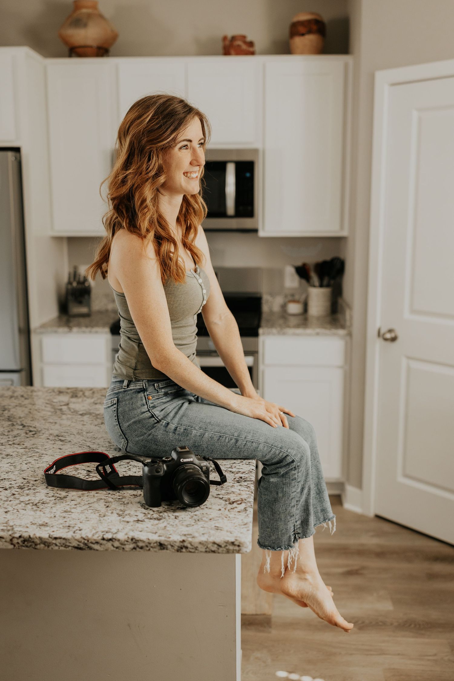 An image showing a lifestyle Woodbury photographer in jeans sitting casually with a camera on a counter in a modern white kitchen.
