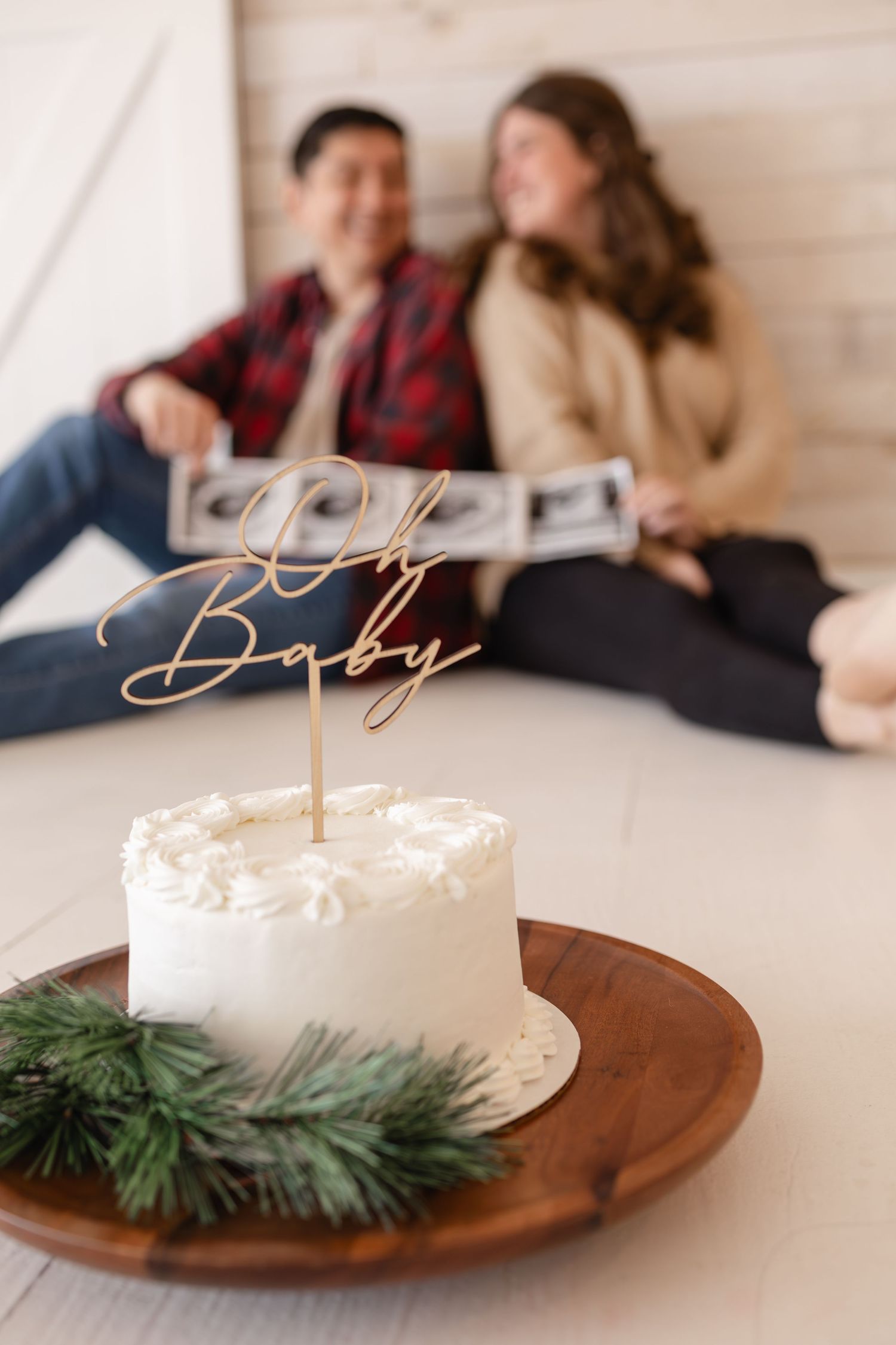 A white cake with gold 'Oh Baby' cake topper and evergreen sprig sits on a wooden serving board taken by Woodbury motherhood photographer.