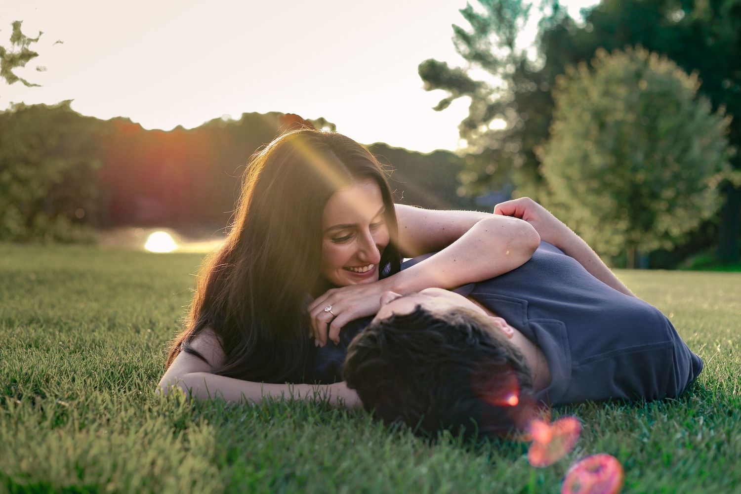 Couple lying together on grass in park during sunset with golden sunlight filtering through trees during Woodbury engagement photos.