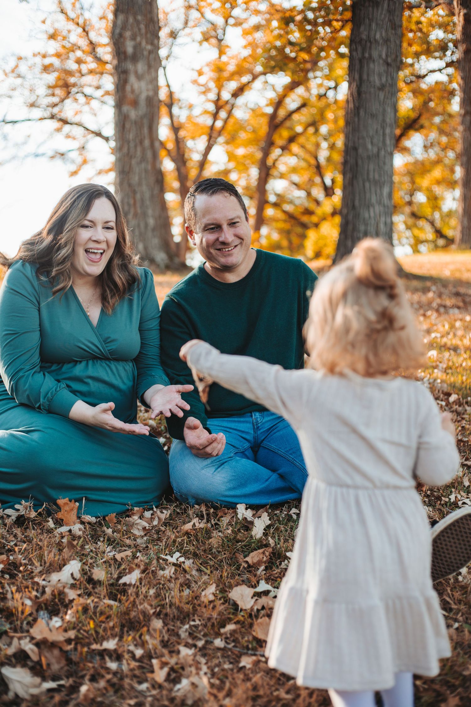 A family sits among autumn leaves in a park, with a toddler girl reaching out to her smiling parents during fall mini session in Lakeville.
