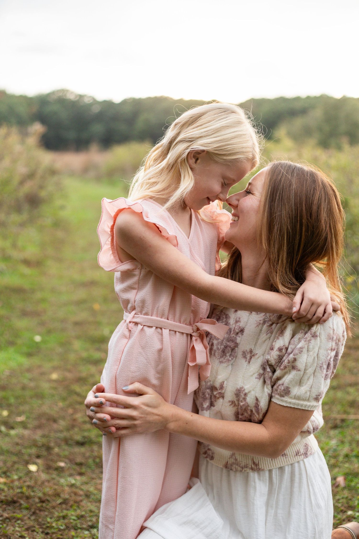 A mother and young daughter share a tender nose-to-nose moment in a field at Tamarack Nature Center, both wearing pink dresses.