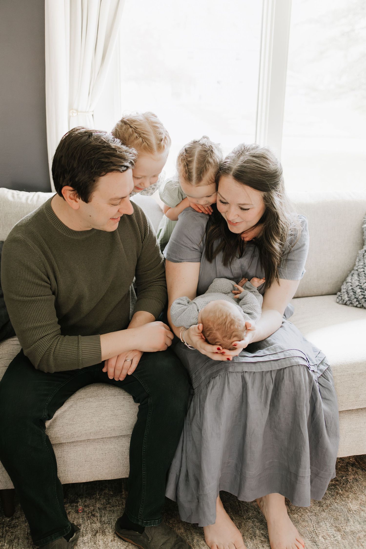 A young family of four sits on a couch, lovingly gazing at their baby wrapped in a gray blanket taken by Woodbury newborn photographer.