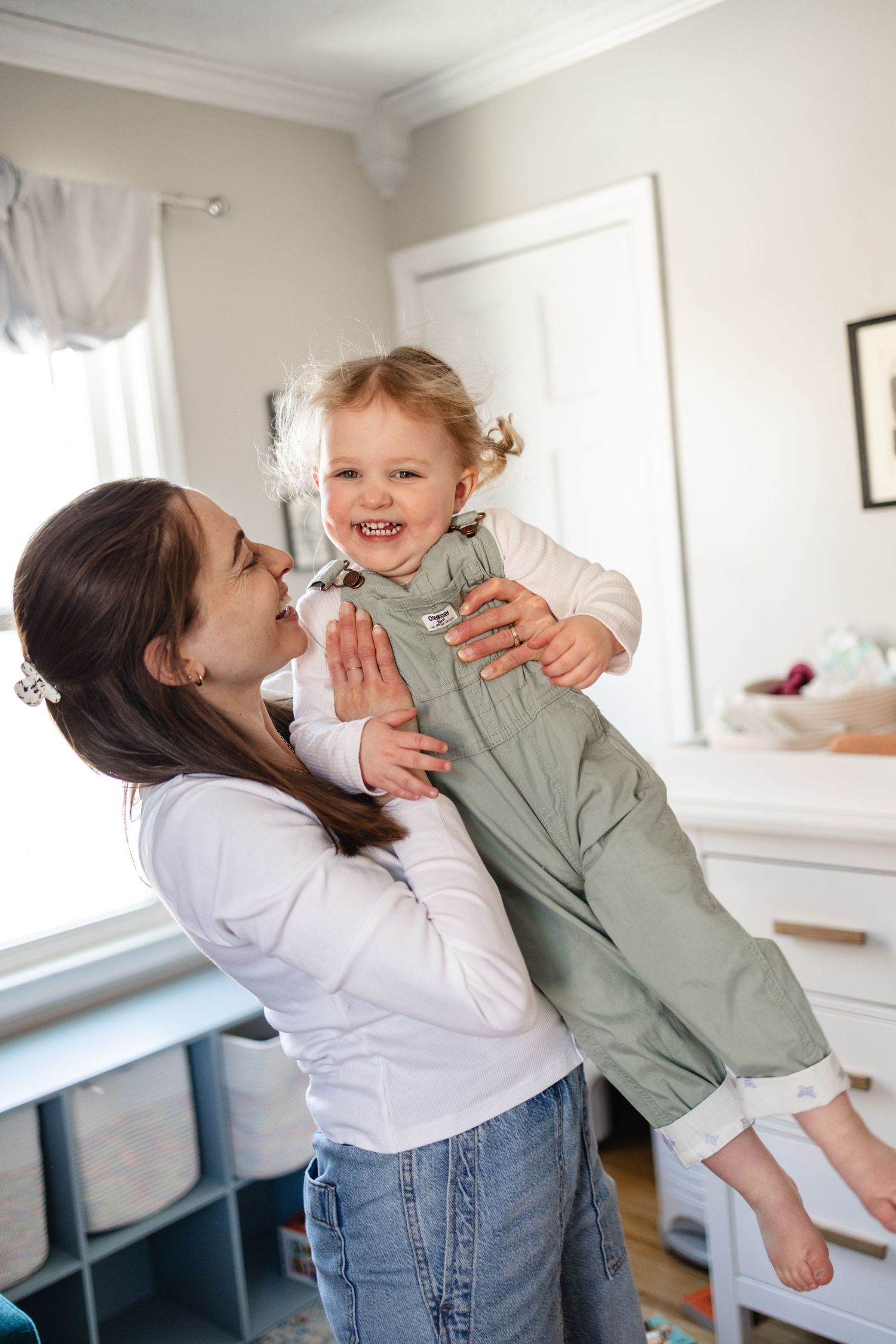 A joyful mother lifts her smiling toddler girl in green overalls in a bright, cozy nursery room in Minneapolis