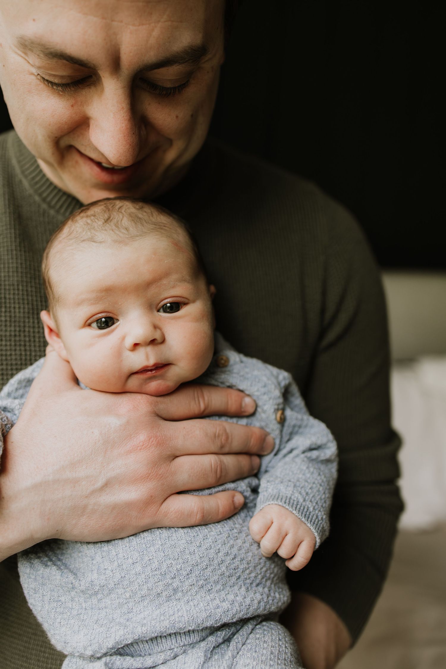 A smiling father gently holds his newborn baby in a cozy gray outfit, sharing a tender moment during in-home newborn session in Twin Cities.