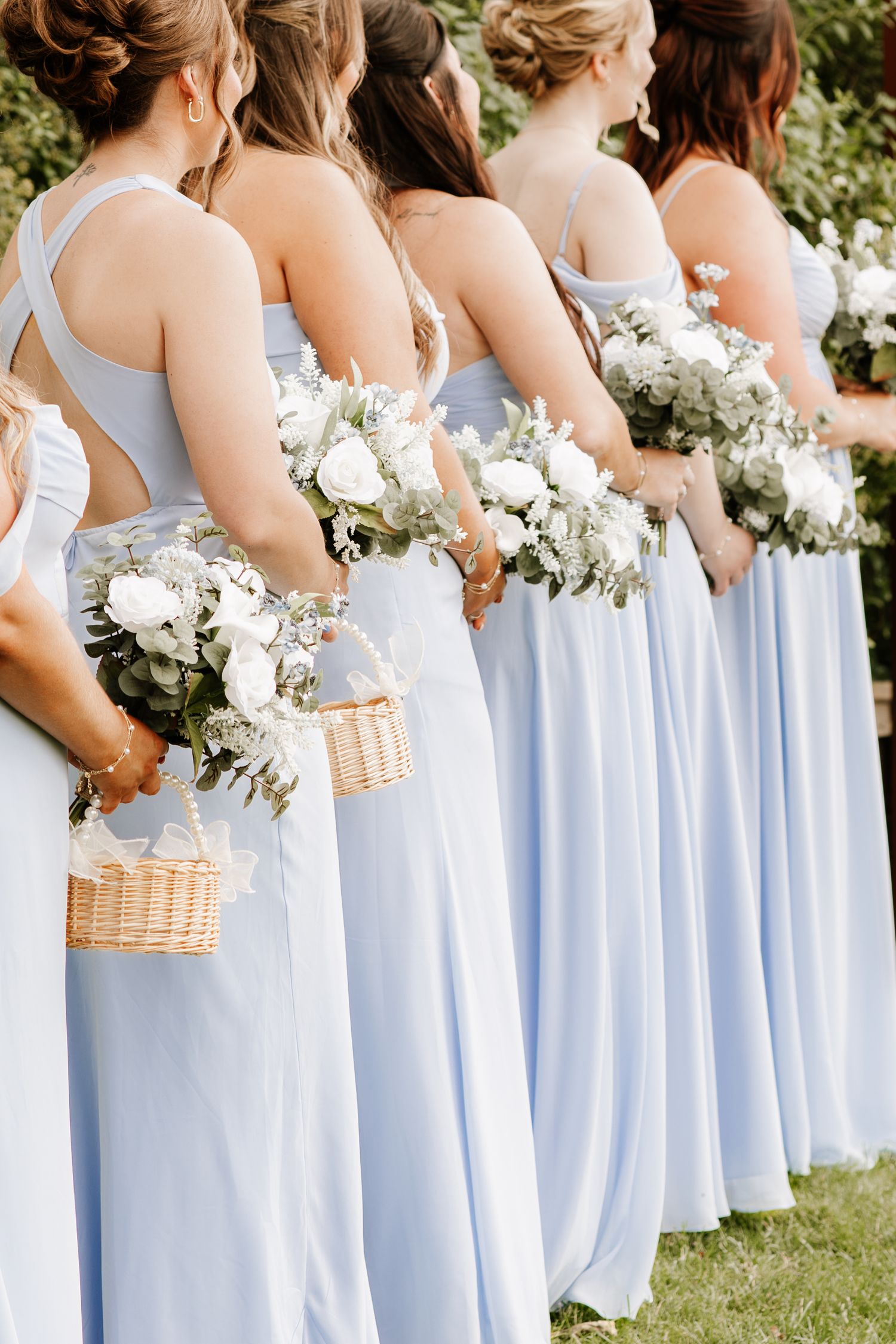 Bridesmaids in light blue gowns holding white floral bouquets lined up outdoors at a wedding ceremony.