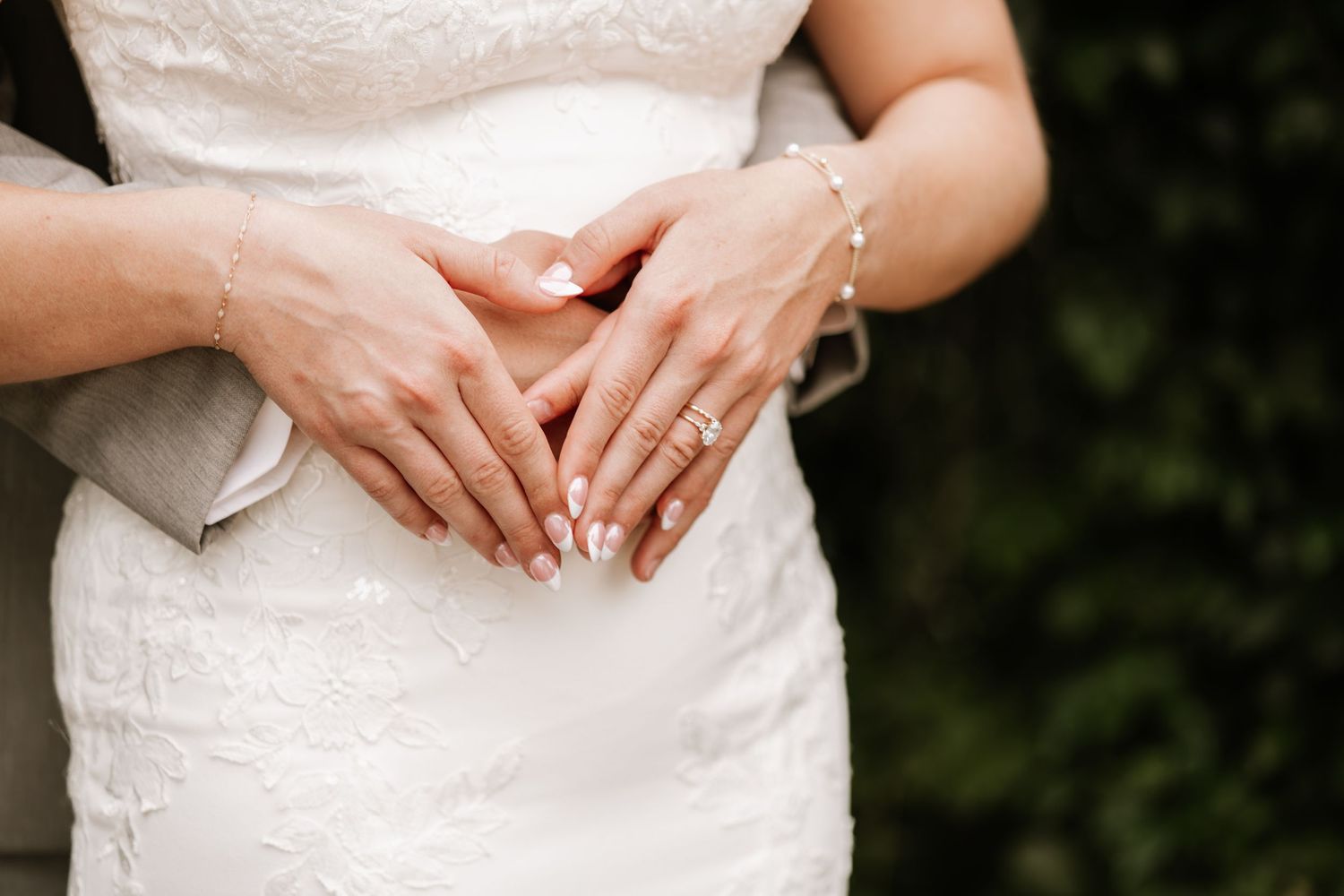 Bride and groom hands forming heart shape on white lace wedding dress, bride wearing diamond ring and bracelet.