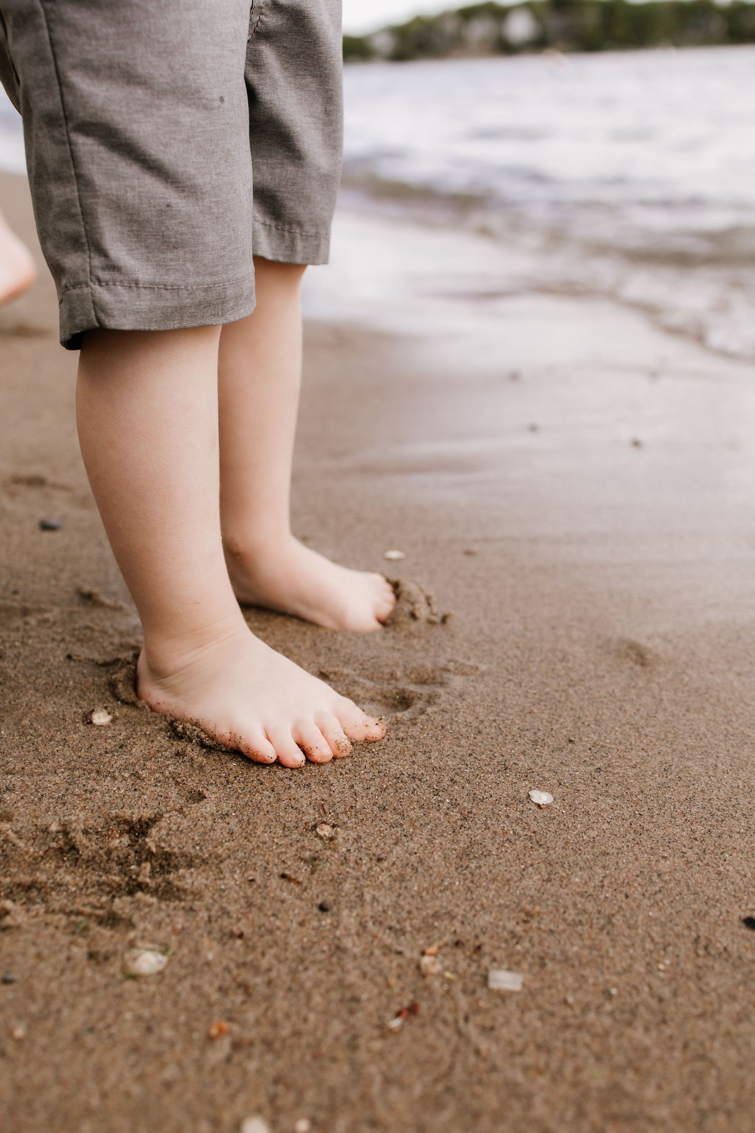 Close-up of a young child's bare feet standing on wet sandy beach shore with gentle ocean waves in the background.